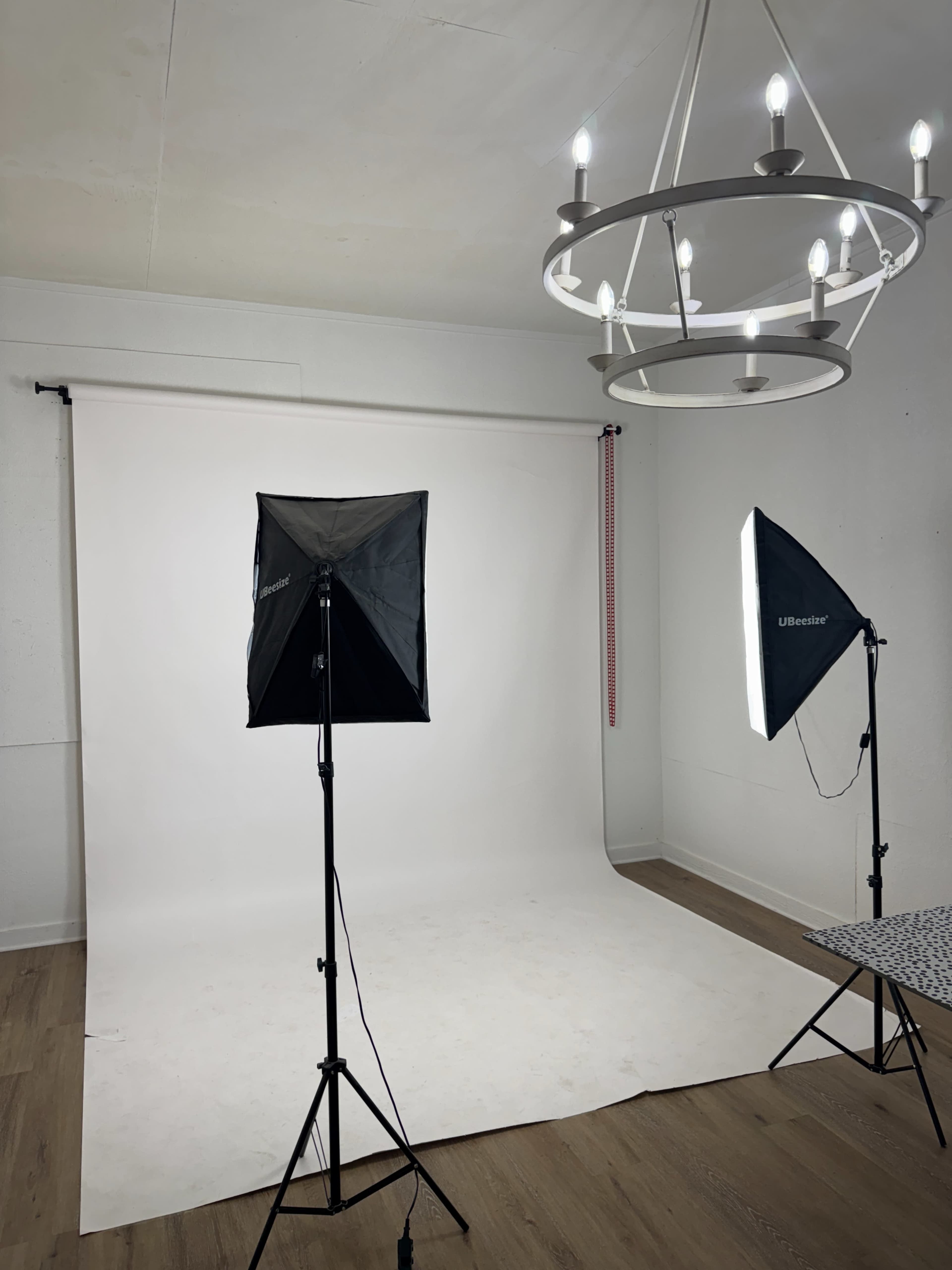 The image shows a photography studio with a white backdrop, two softbox lights positioned on either side, and a chandelier hanging from the ceiling.