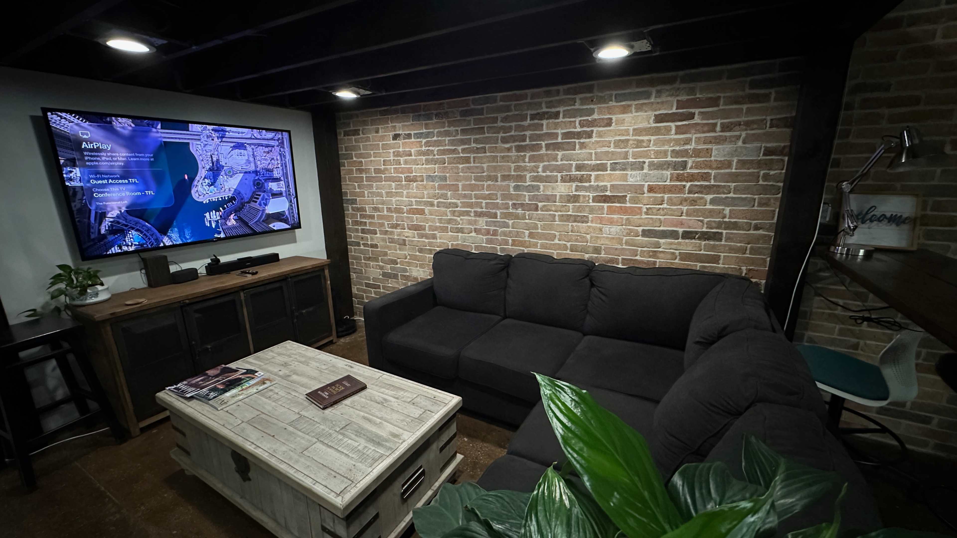 The image shows a cozy living area with a dark sectional couch, a rustic coffee table, and a television mounted on a brick wall, illuminated by overhead lights.