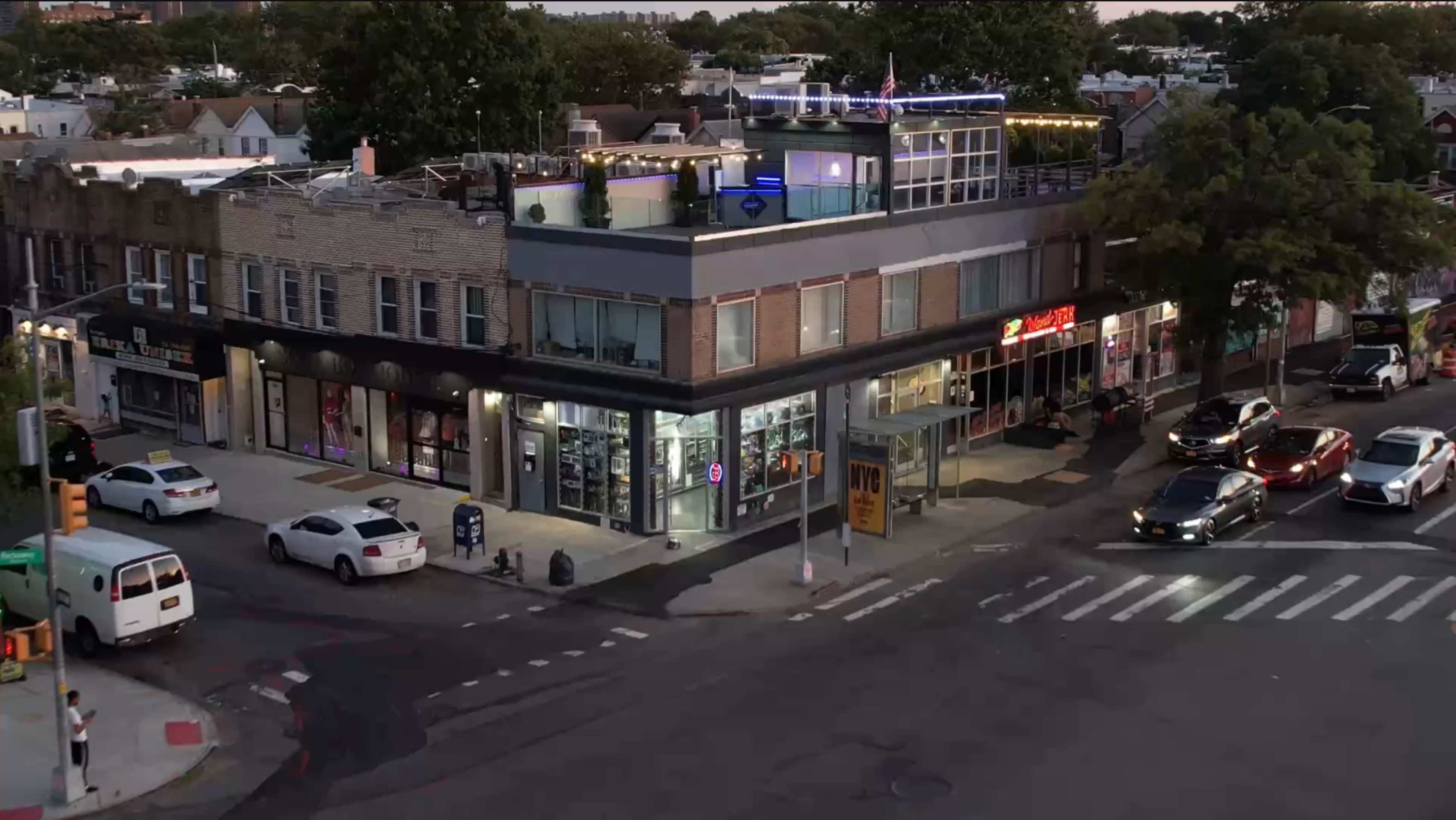 A corner building features a rooftop terrace and various businesses on the ground floor, with cars parked and moving along the intersection.