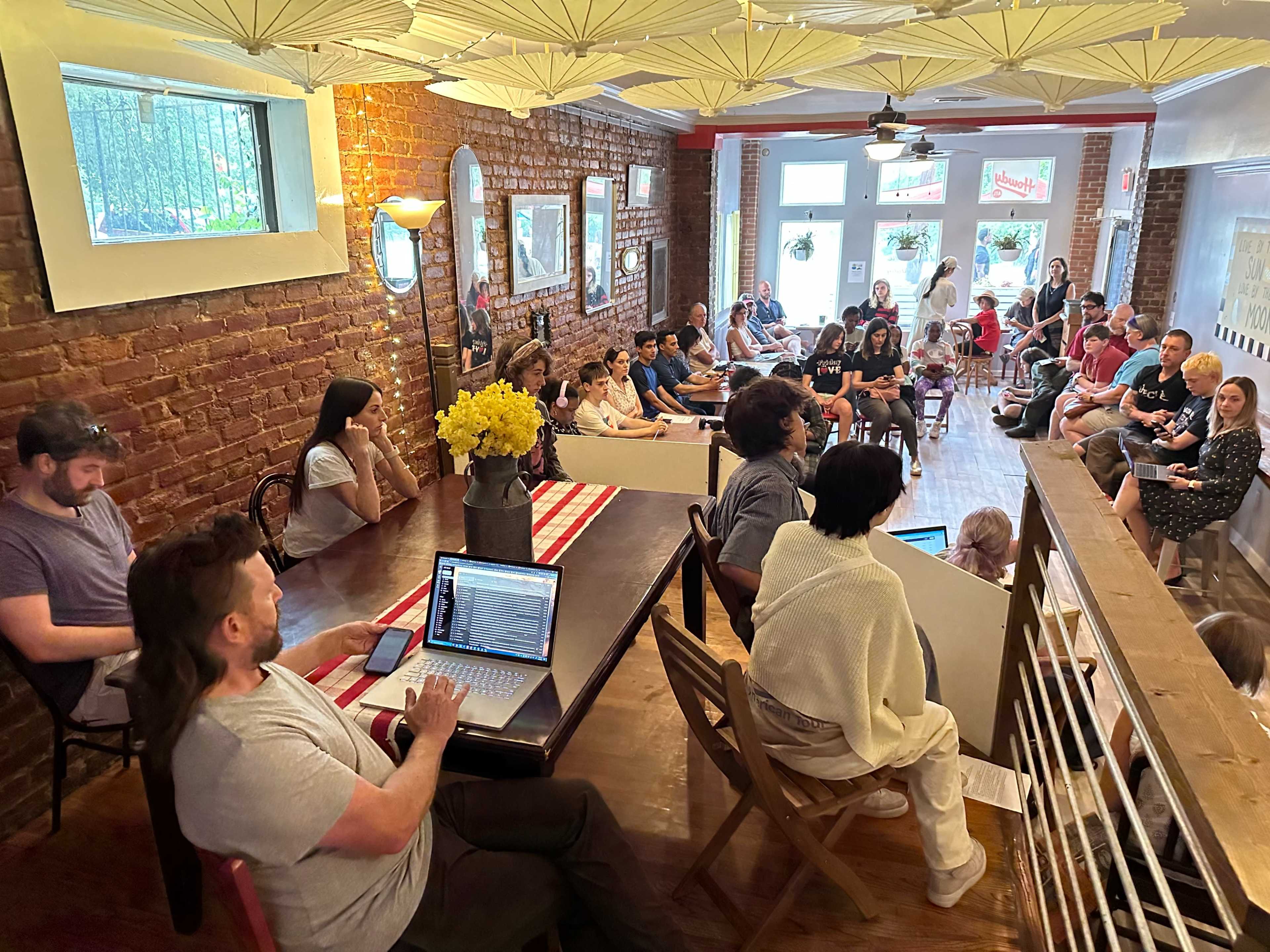 A group of people are gathered in a cozy room with exposed brick walls for a discussion or presentation, while others sit attentively in chairs.
