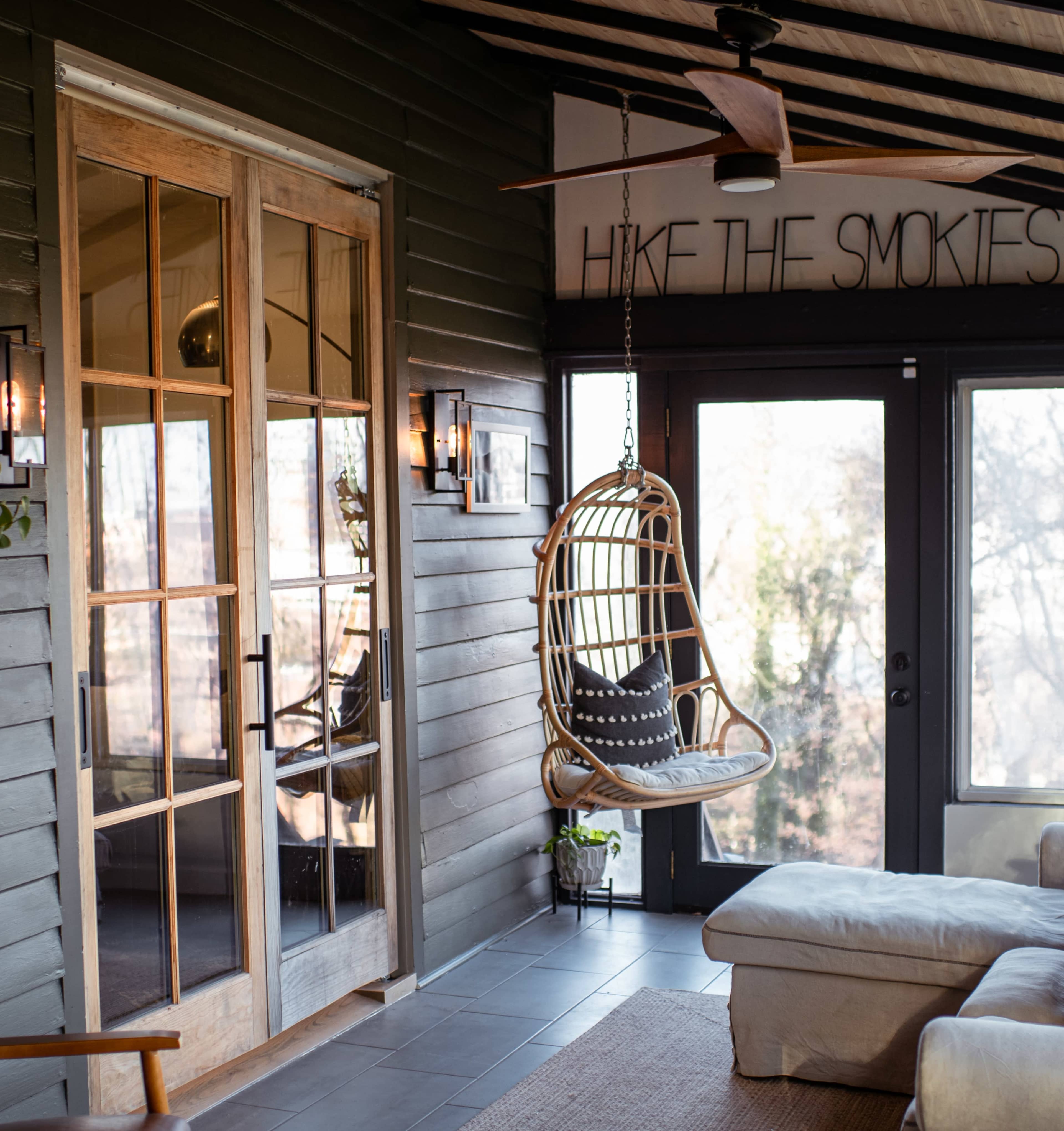 A cozy indoor space featuring a hanging wicker chair, large glass doors, and a wall sign that reads "HIKE THE SMOKIES."