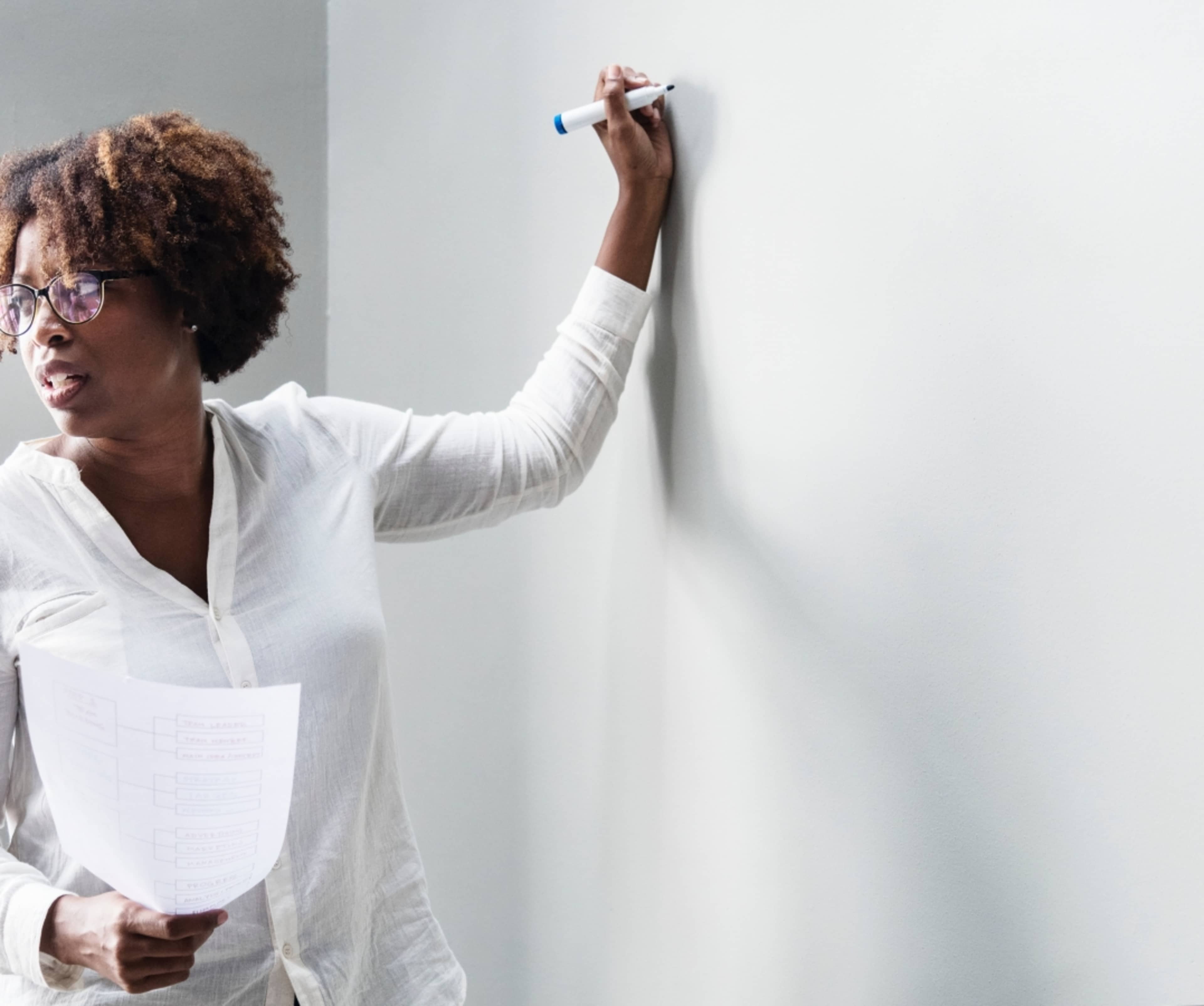 A woman with curly hair is writing on a blank wall while holding a printed document in her other hand.