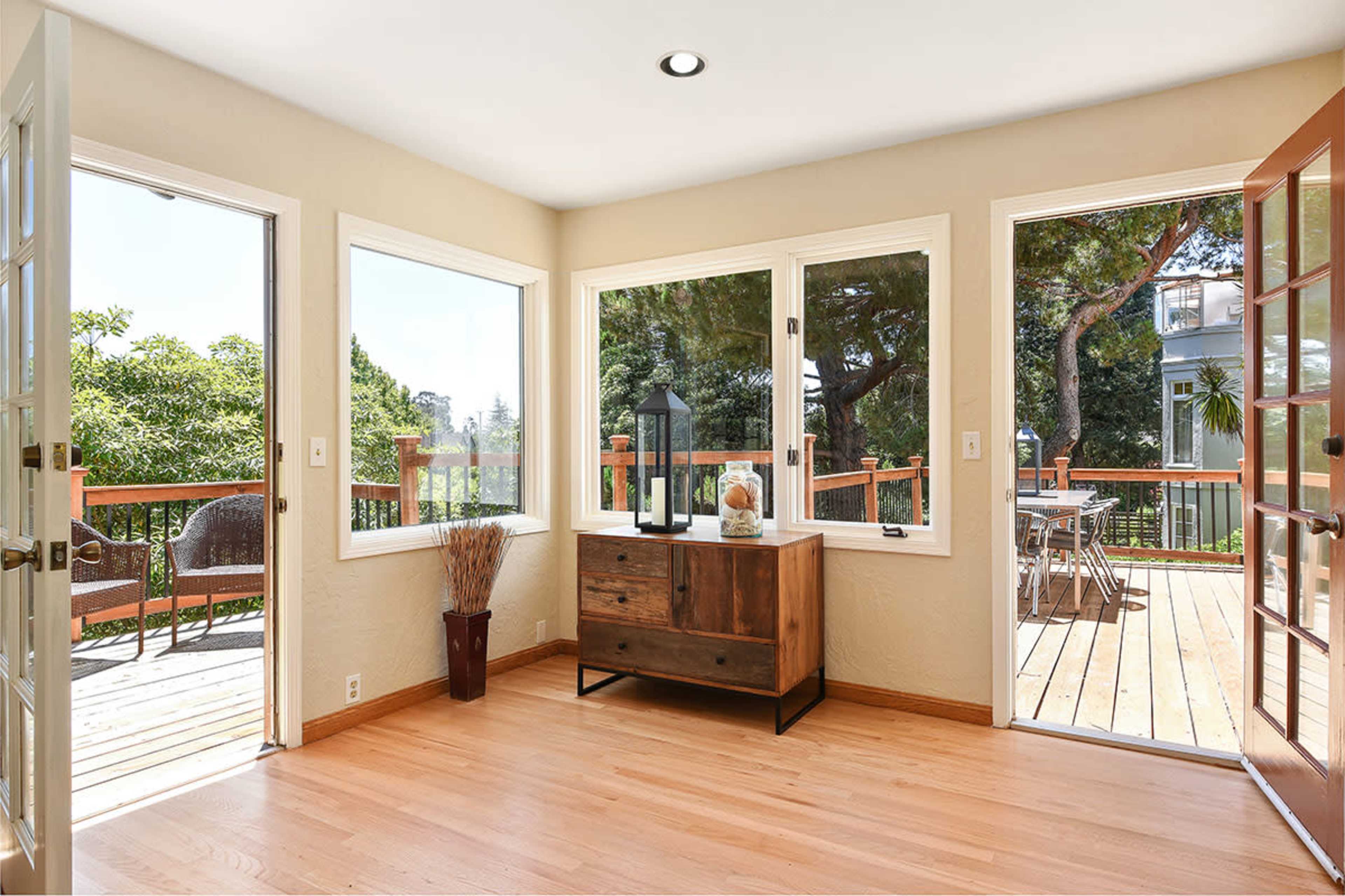 The room features large windows that provide ample natural light, a wooden sideboard against the wall, and a view of a wooden deck surrounded by greenery.