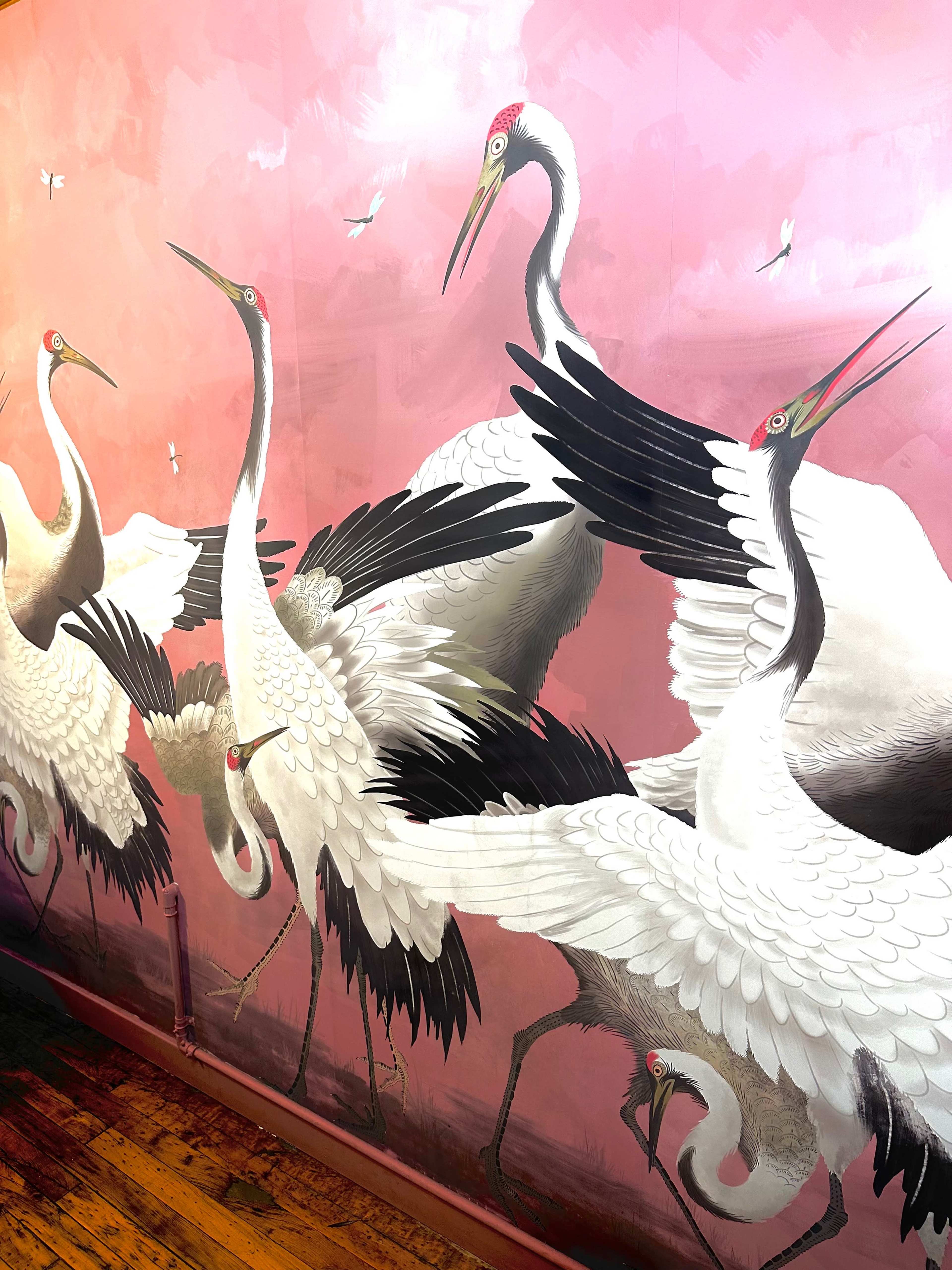 A vibrant mural depicting several cranes with striking black and white plumage against a pink backdrop.