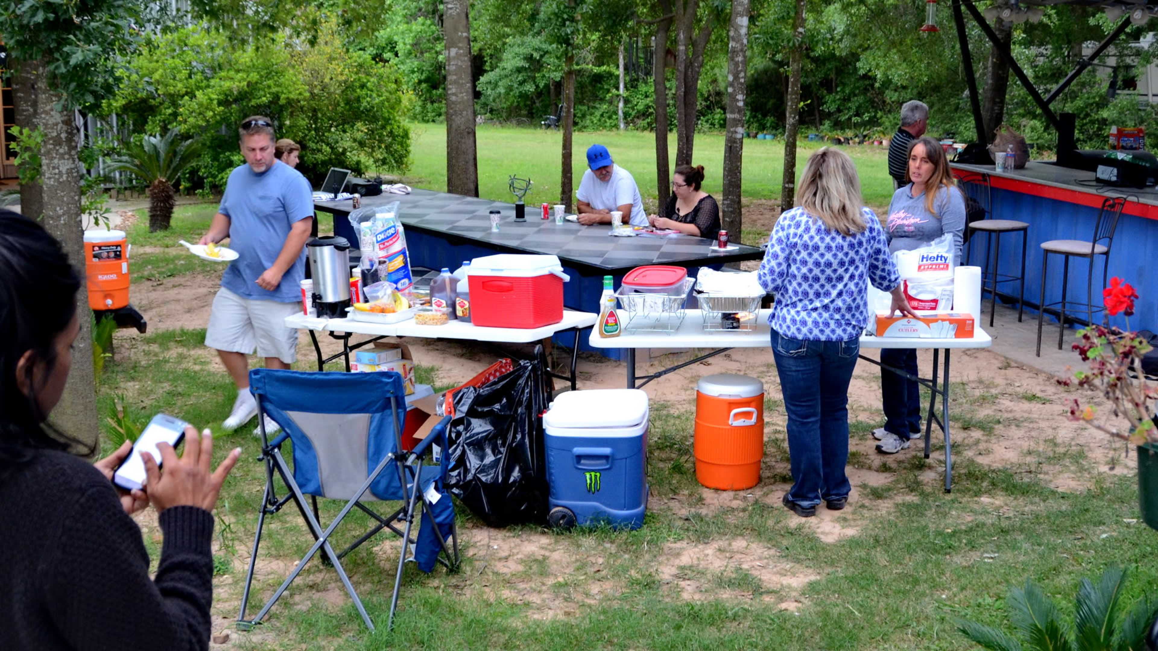 A group of people gathers in a backyard for a barbecue, with food laid out on tables and others seated nearby.