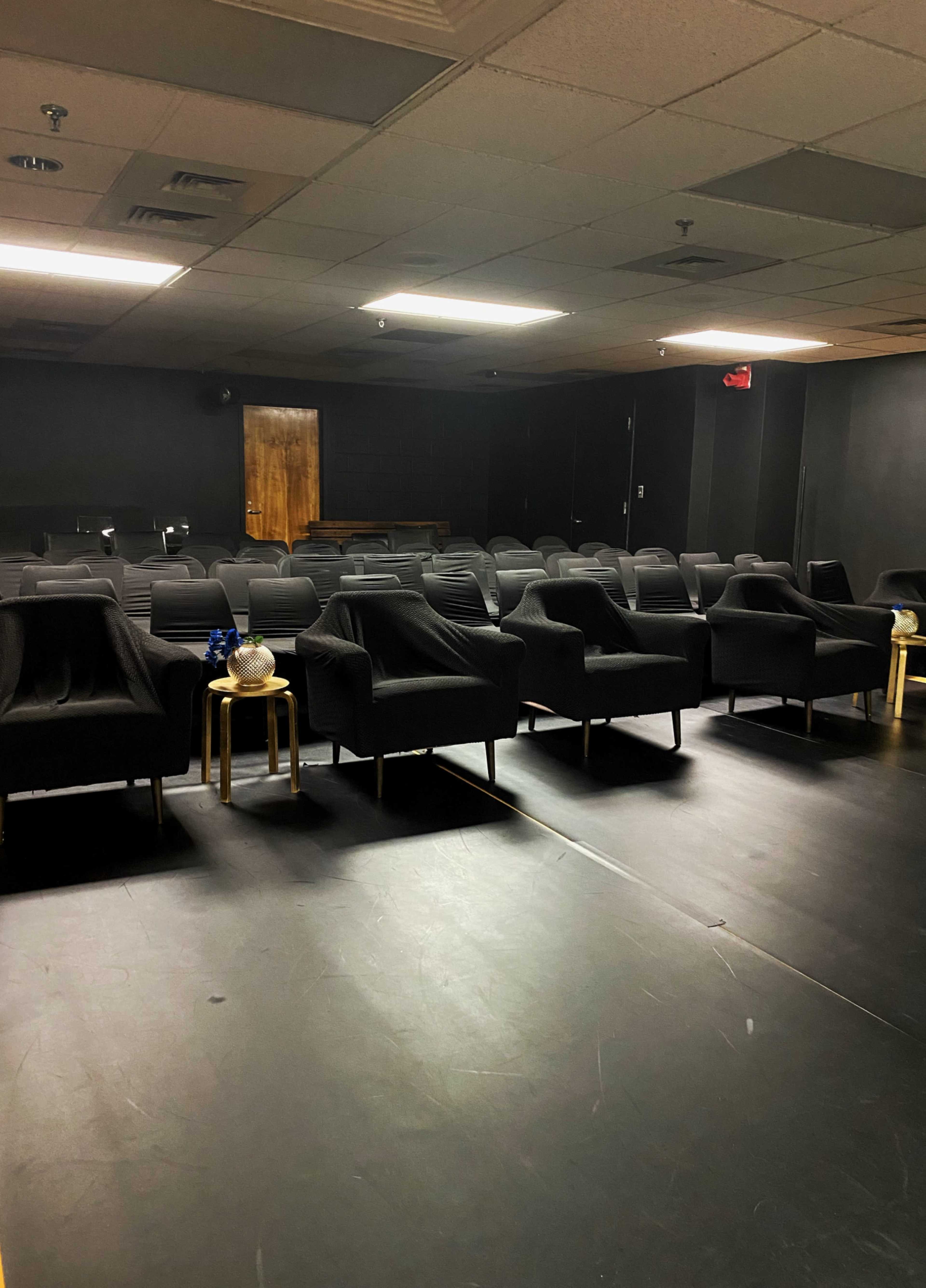 A dimly lit room features rows of black chairs and small tables set against a dark backdrop.