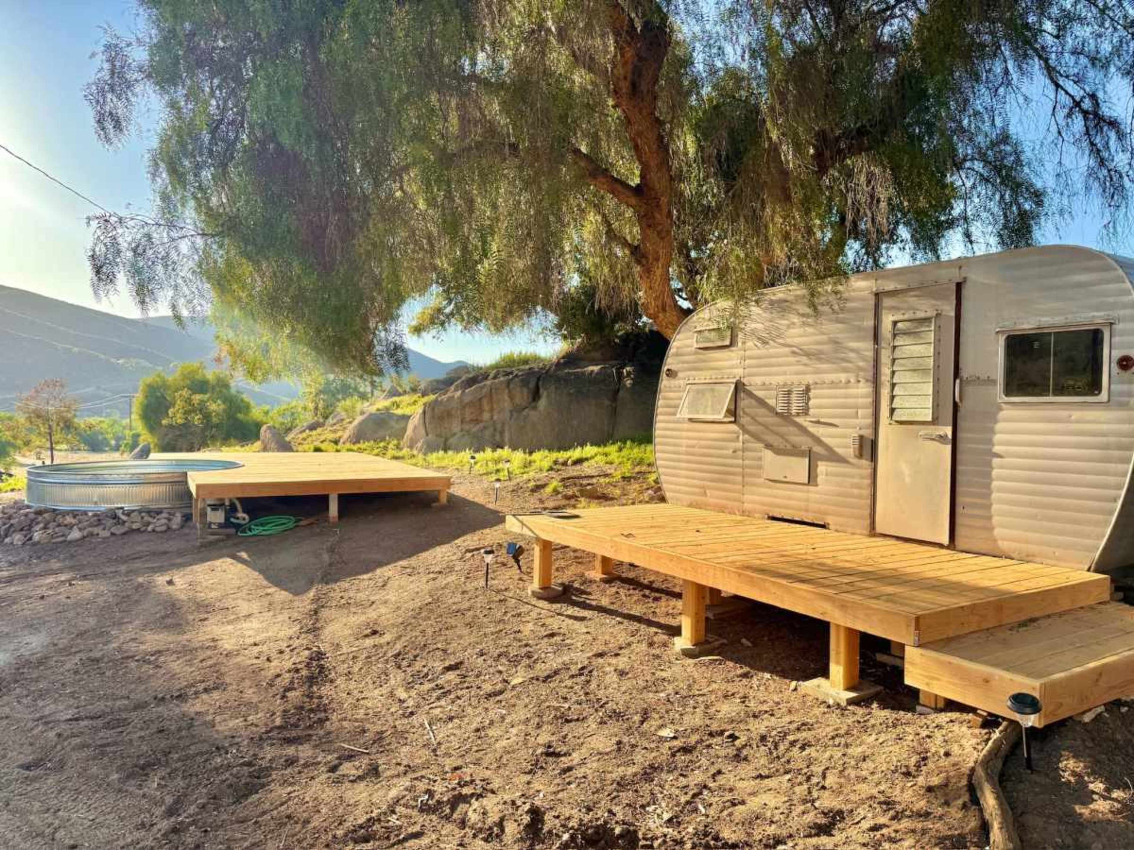 A silver vintage trailer sits beside wooden decks and a round metal fire pit under a large tree with mountains in the background.