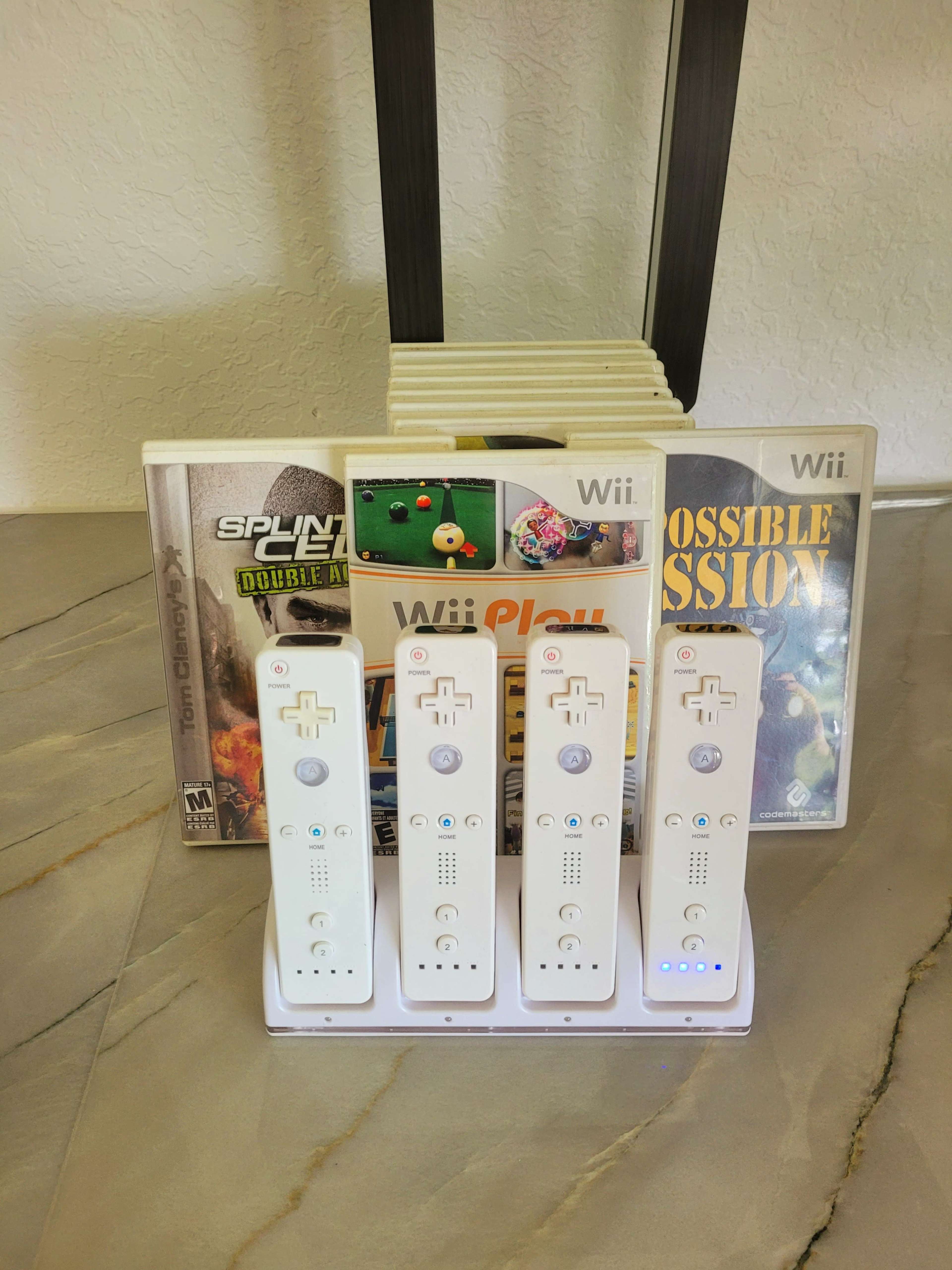 The image shows several Wii controllers neatly placed on a charging dock alongside a stack of Wii game cases including "Splinter Cell: Double Agent" and "Impossible Mission."