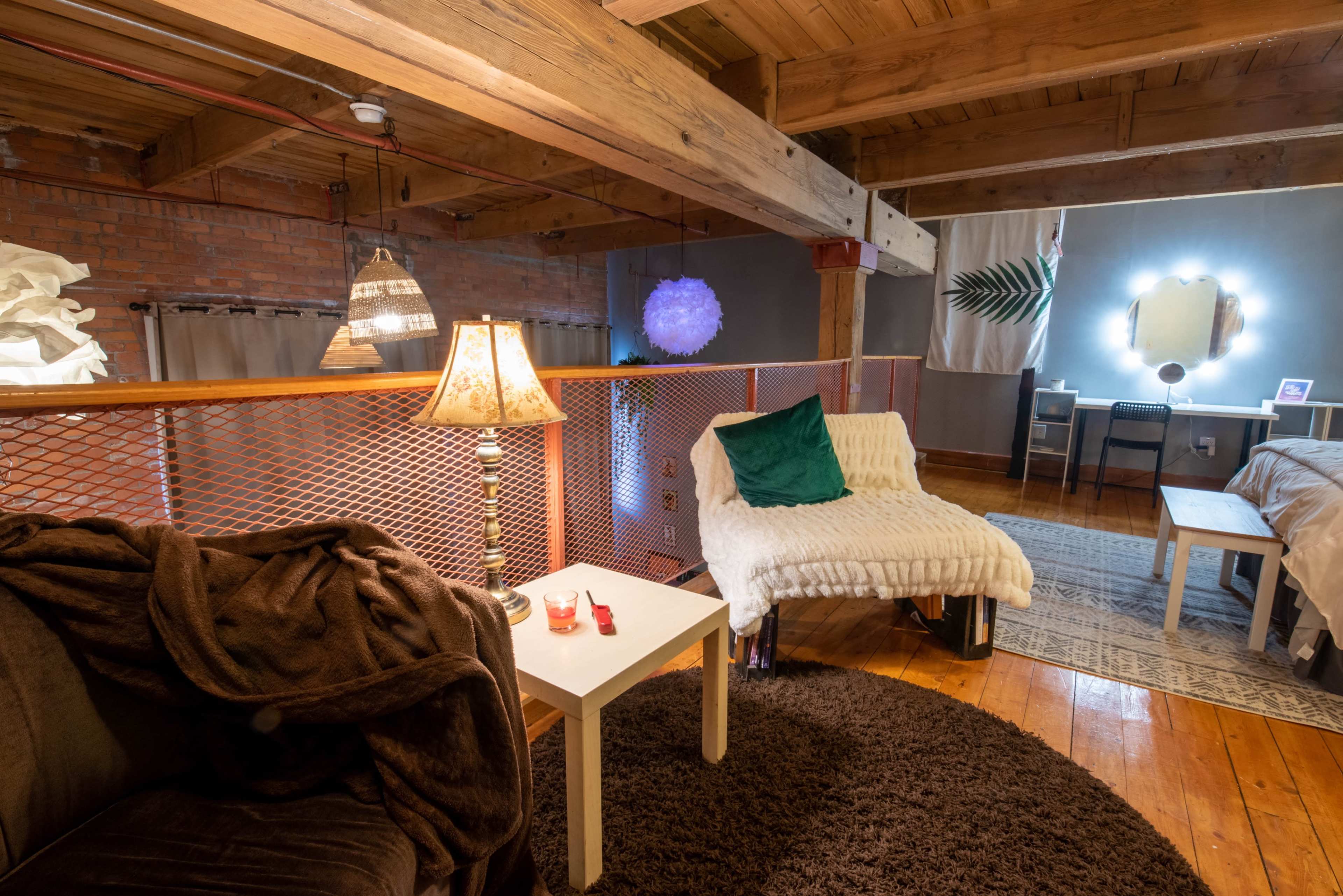 A cozy loft space features a seating area with a brown blanket draped on a couch, a small table with candles, and a work desk illuminated by a mirror with lights.