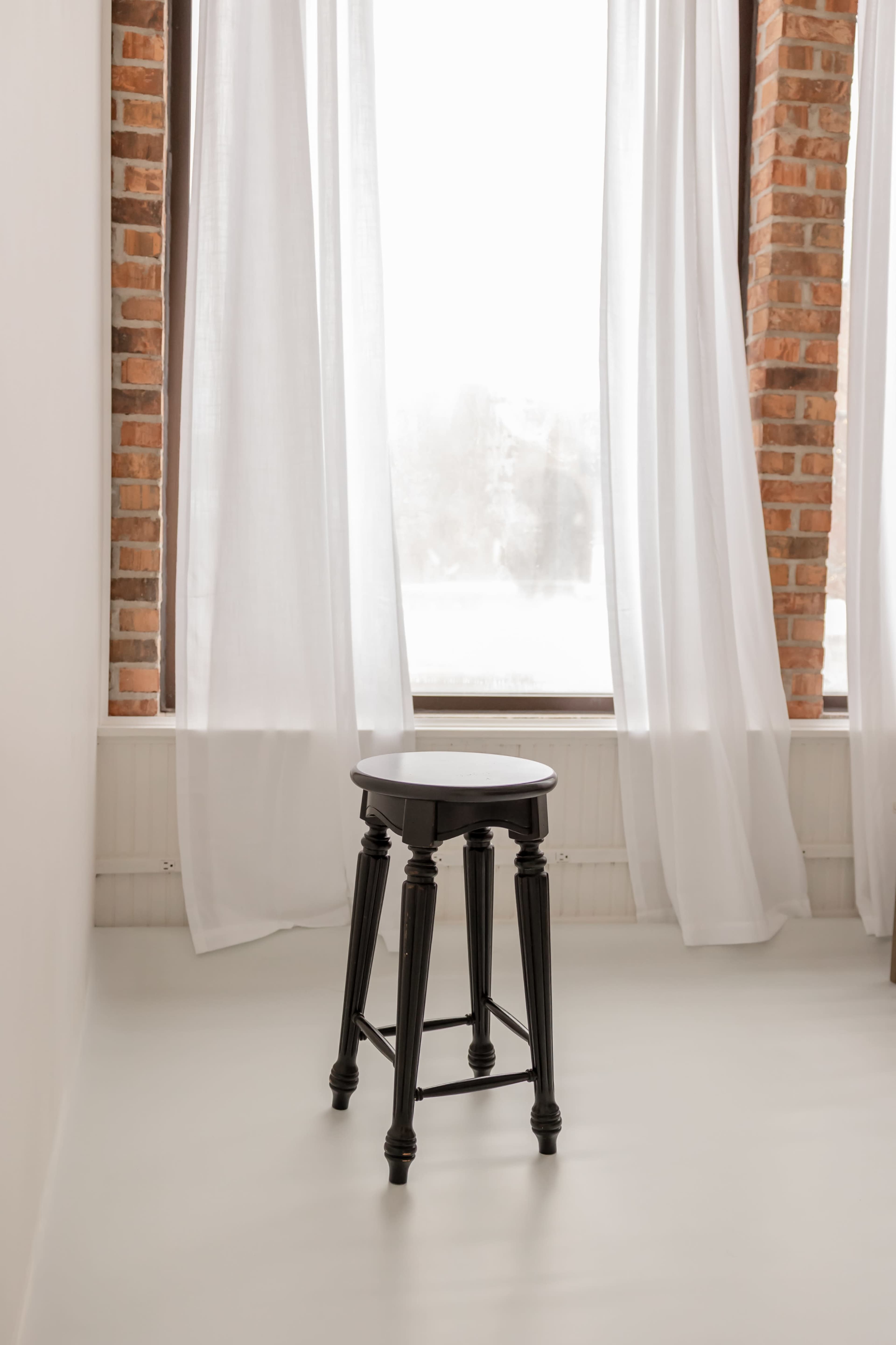 A black wooden stool stands alone near a window draped with sheer white curtains, in a room with brick walls.