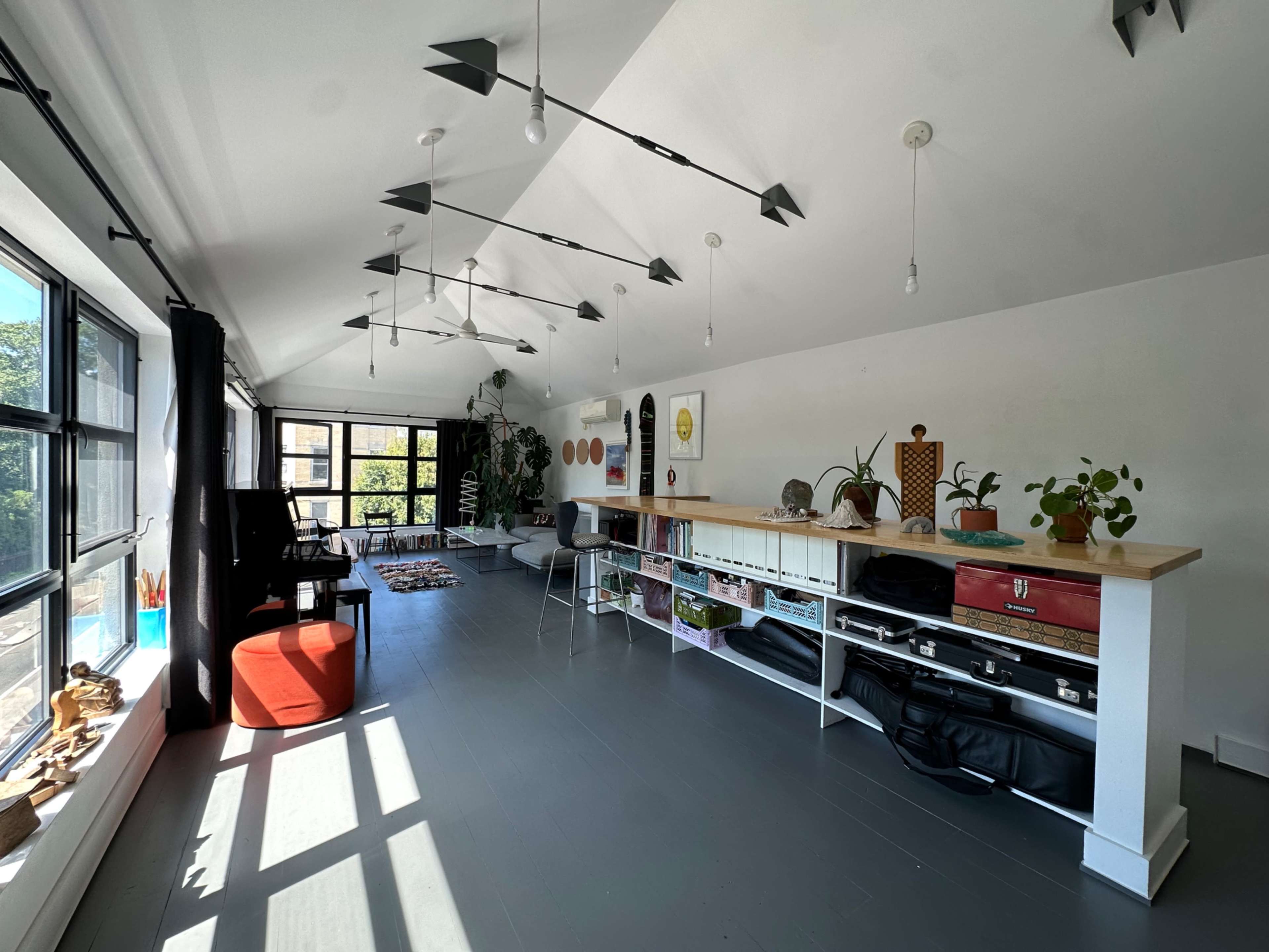 The image depicts a bright, spacious room with large windows, modern furniture, and shelving displaying various plants and decorative items.