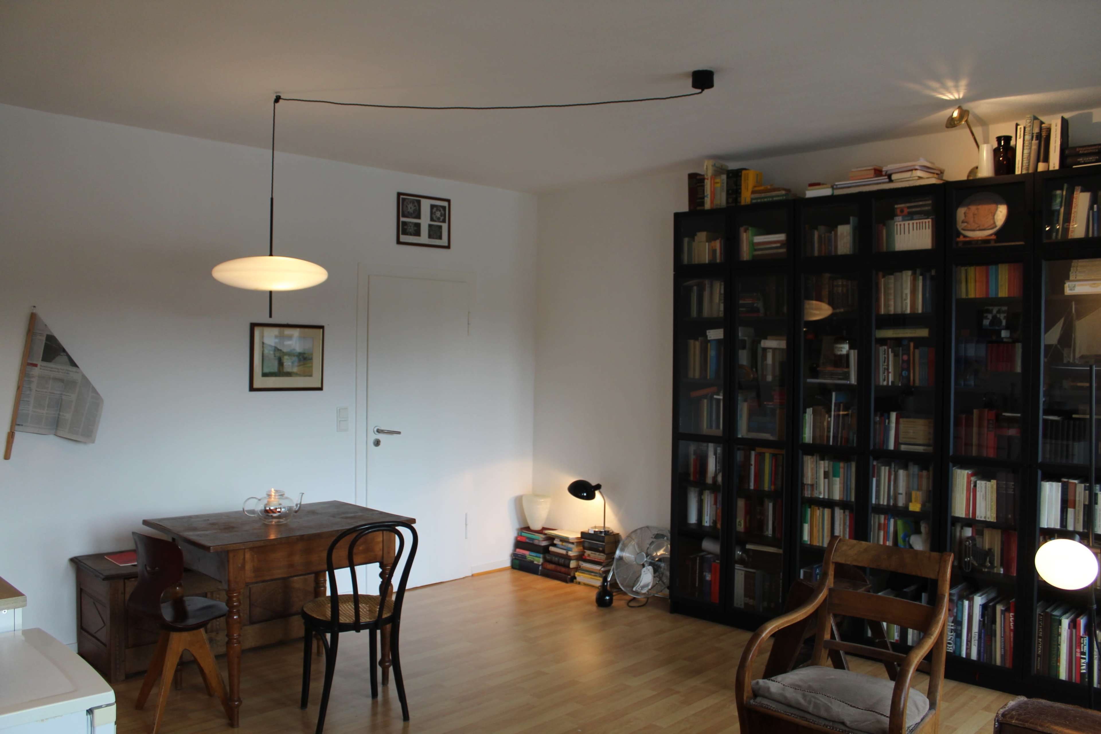 A minimalist room features a wooden desk and chair, a large bookshelf filled with books, and a floor lamp, with a light fixture hanging from the ceiling.