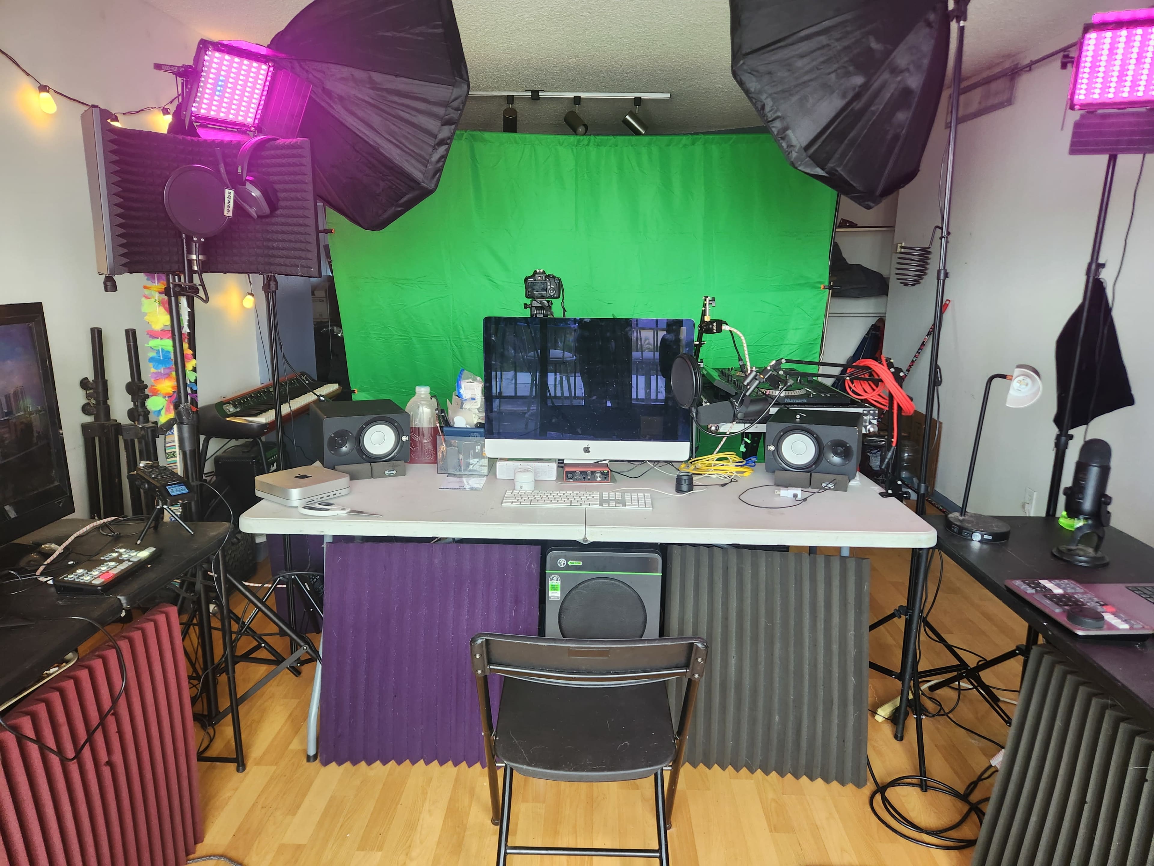 The image shows a recording studio setup with a large green backdrop, multiple studio lights, and various audio equipment arranged on a table.