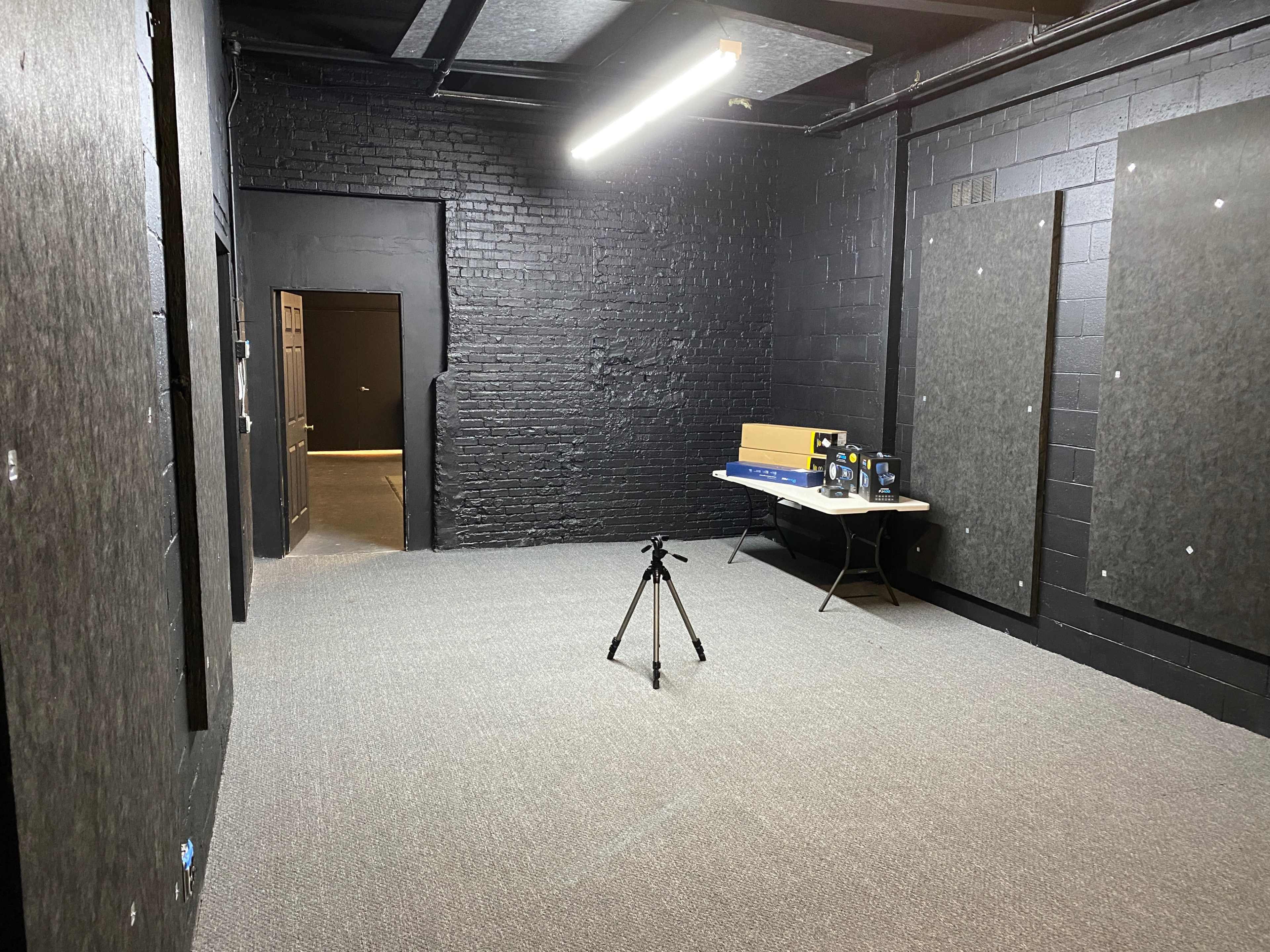 A dark room with black walls, a gray carpet, a table with equipment on it, and a camera tripod set up in the center.