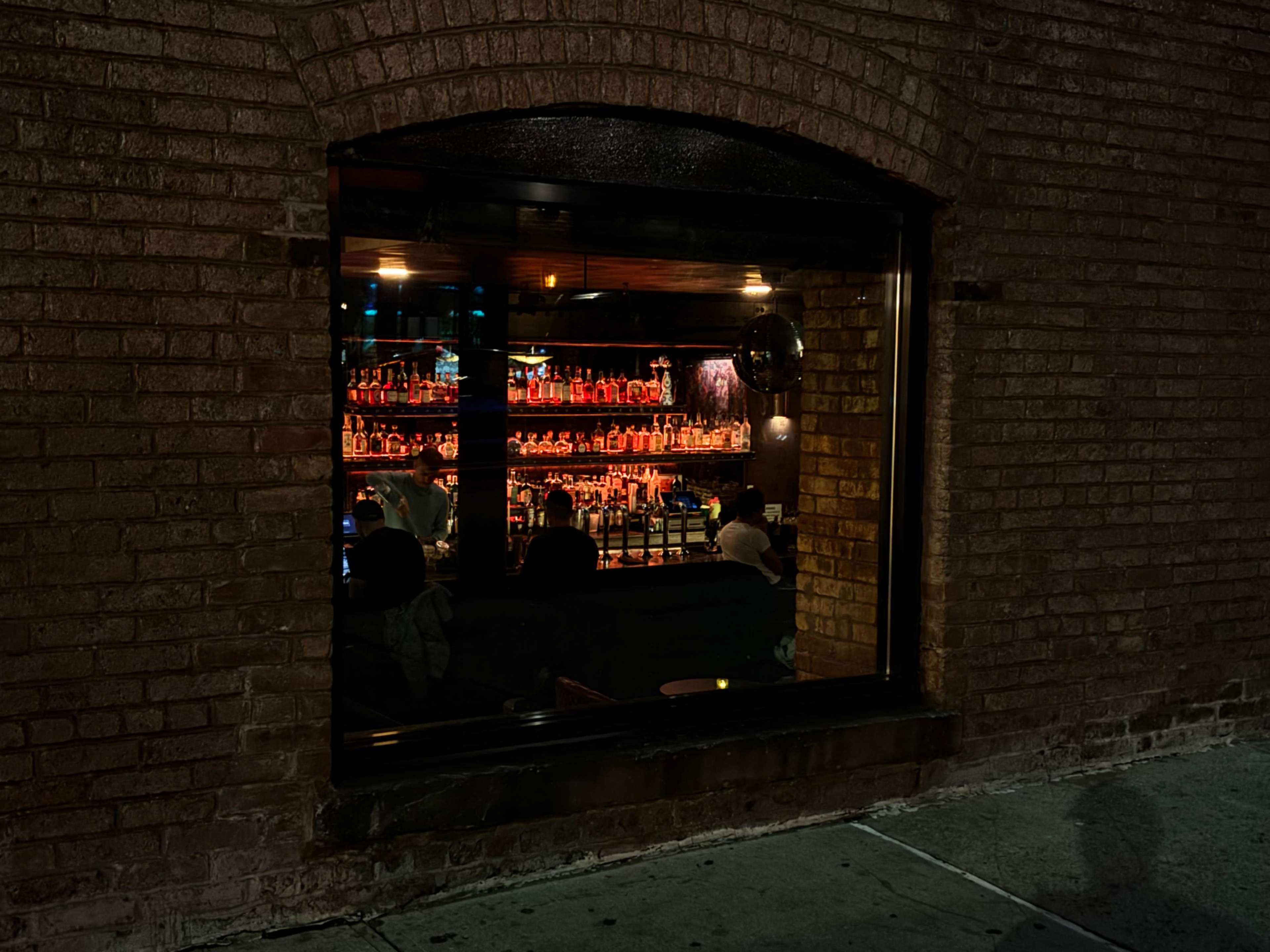 A brightly lit bar filled with colorful liquor bottles is visible through a large window in a brick wall.