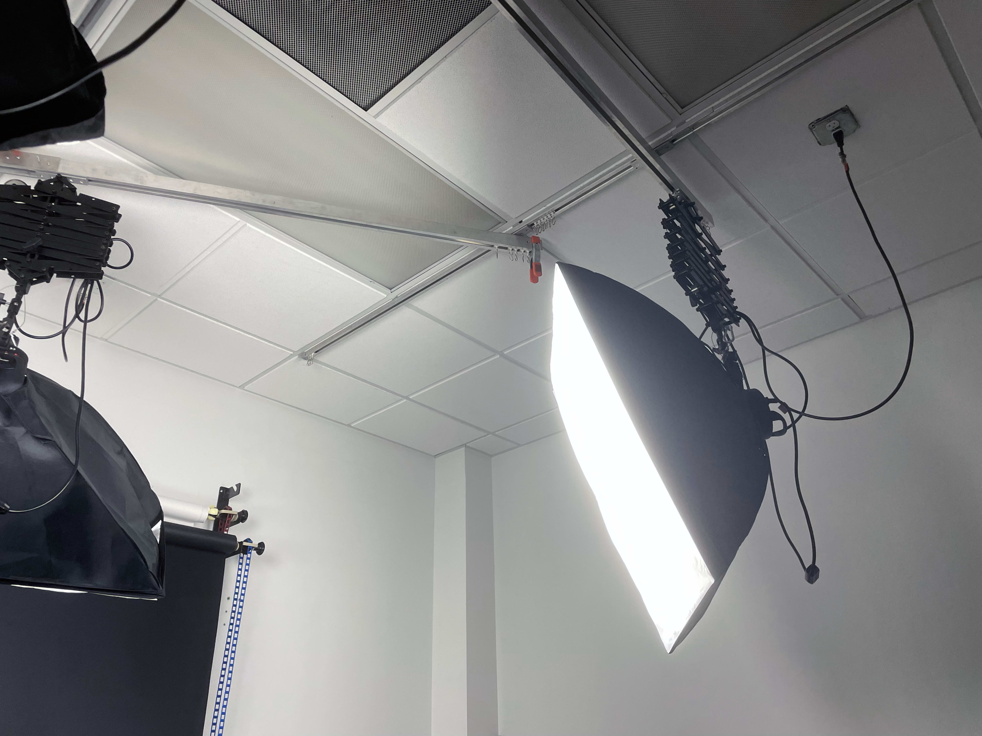 A photography studio setup with softbox lights mounted on the ceiling and a plain white wall in the background.