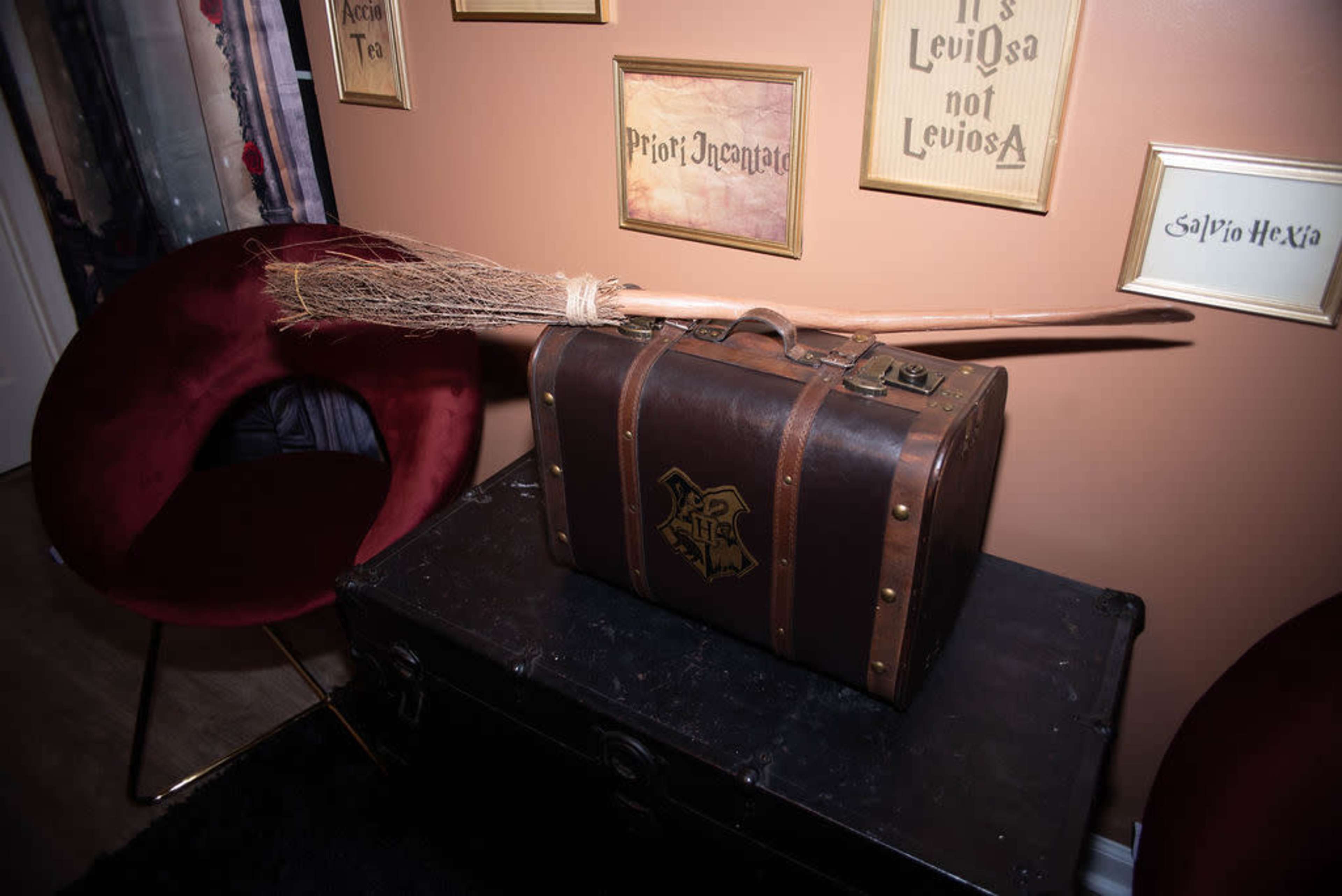 A brown leather suitcase with a broom resting on top is placed beside a red chair against a beige wall adorned with framed signs.