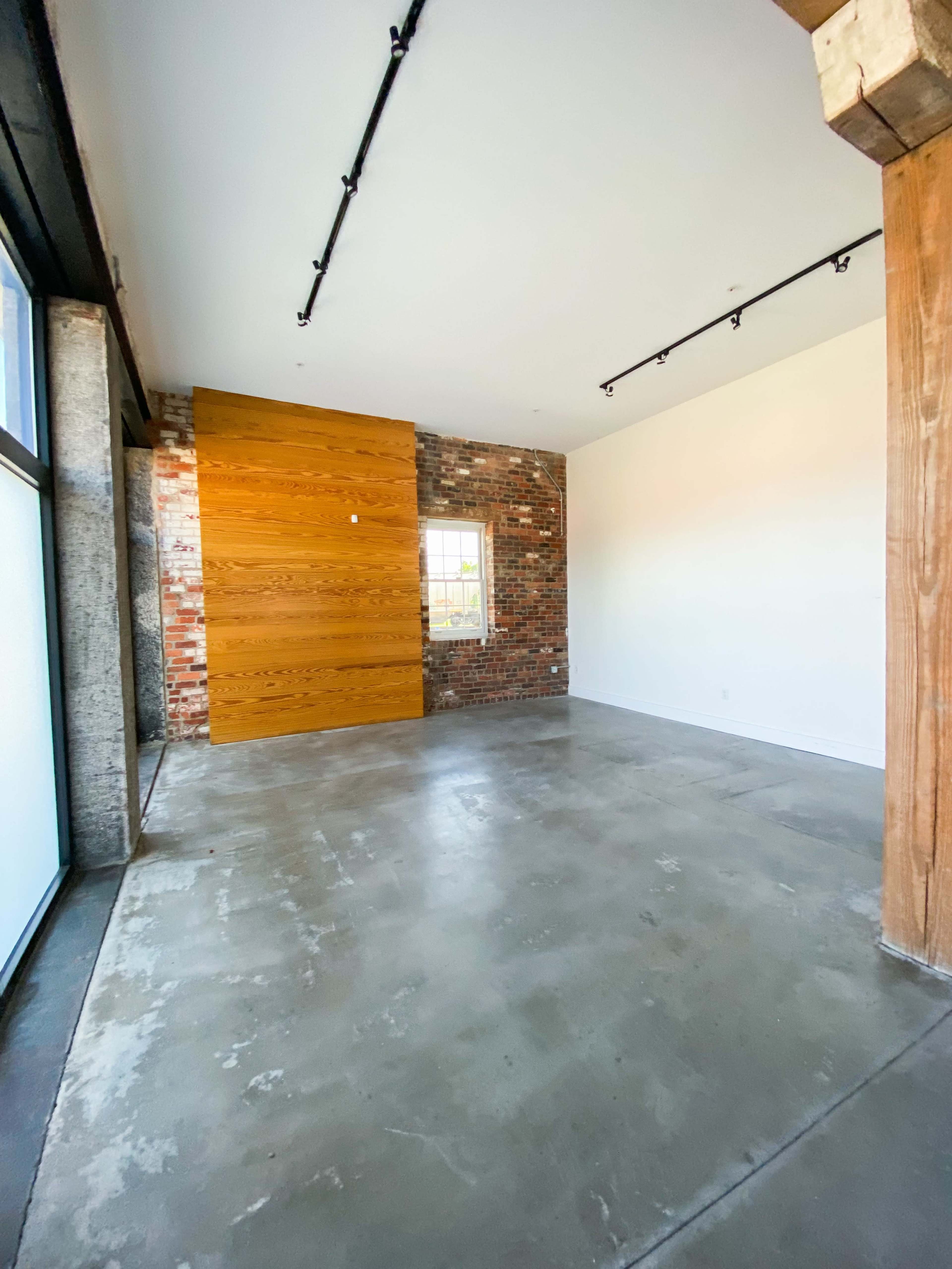 The image shows a spacious room featuring a mixture of exposed brick and wooden wall accents, with concrete flooring and large windows.