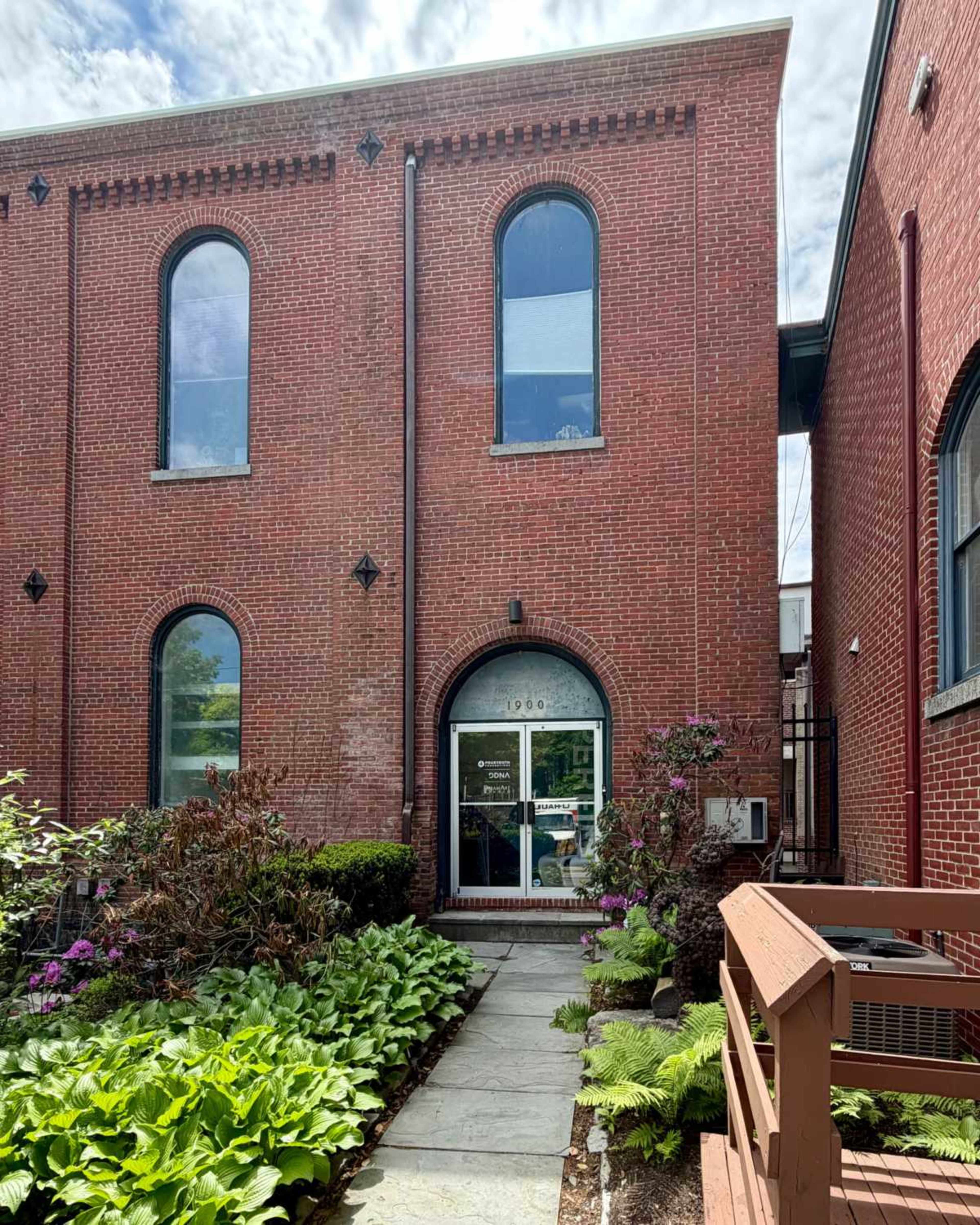 The image shows a red brick building with arched windows and a landscaped entrance featuring green plants and wooden steps.
