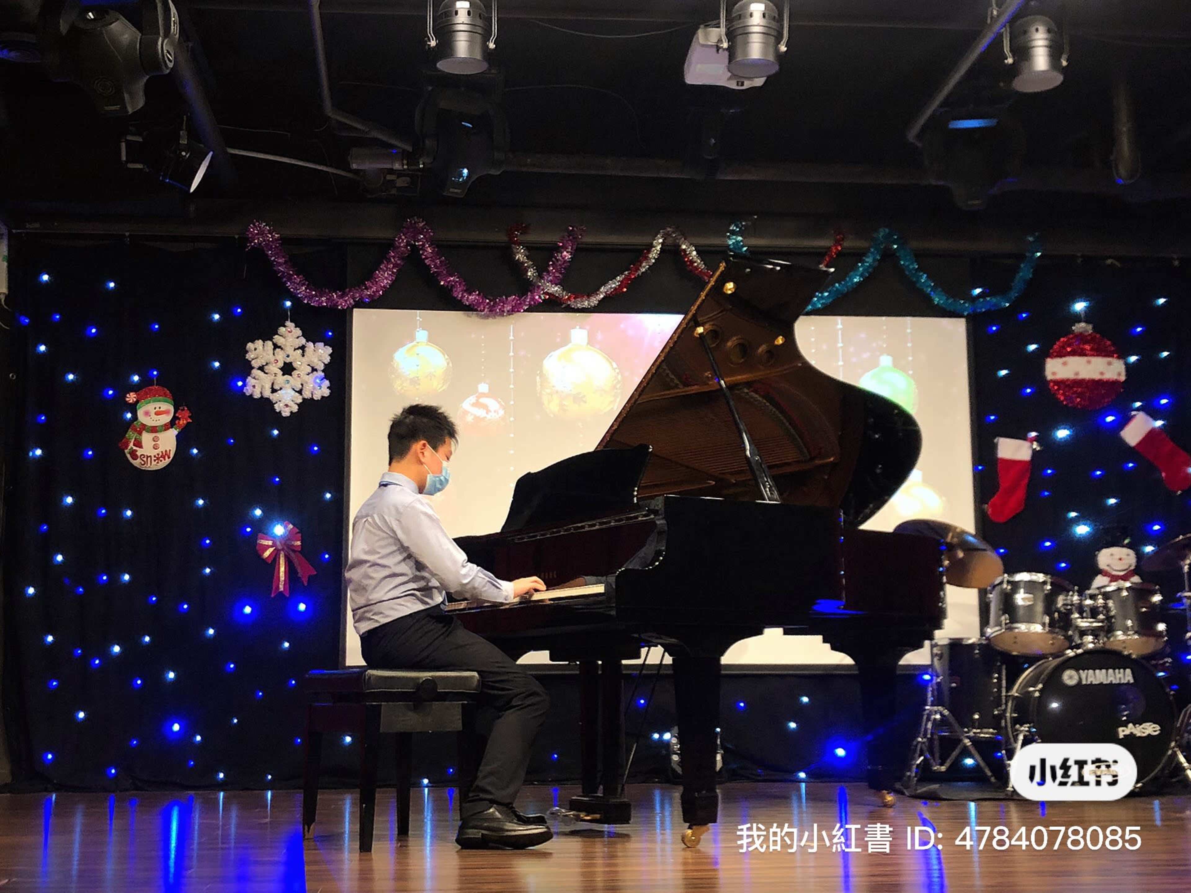 A person in a mask plays a grand piano on stage with decorative lights and a festive backdrop.