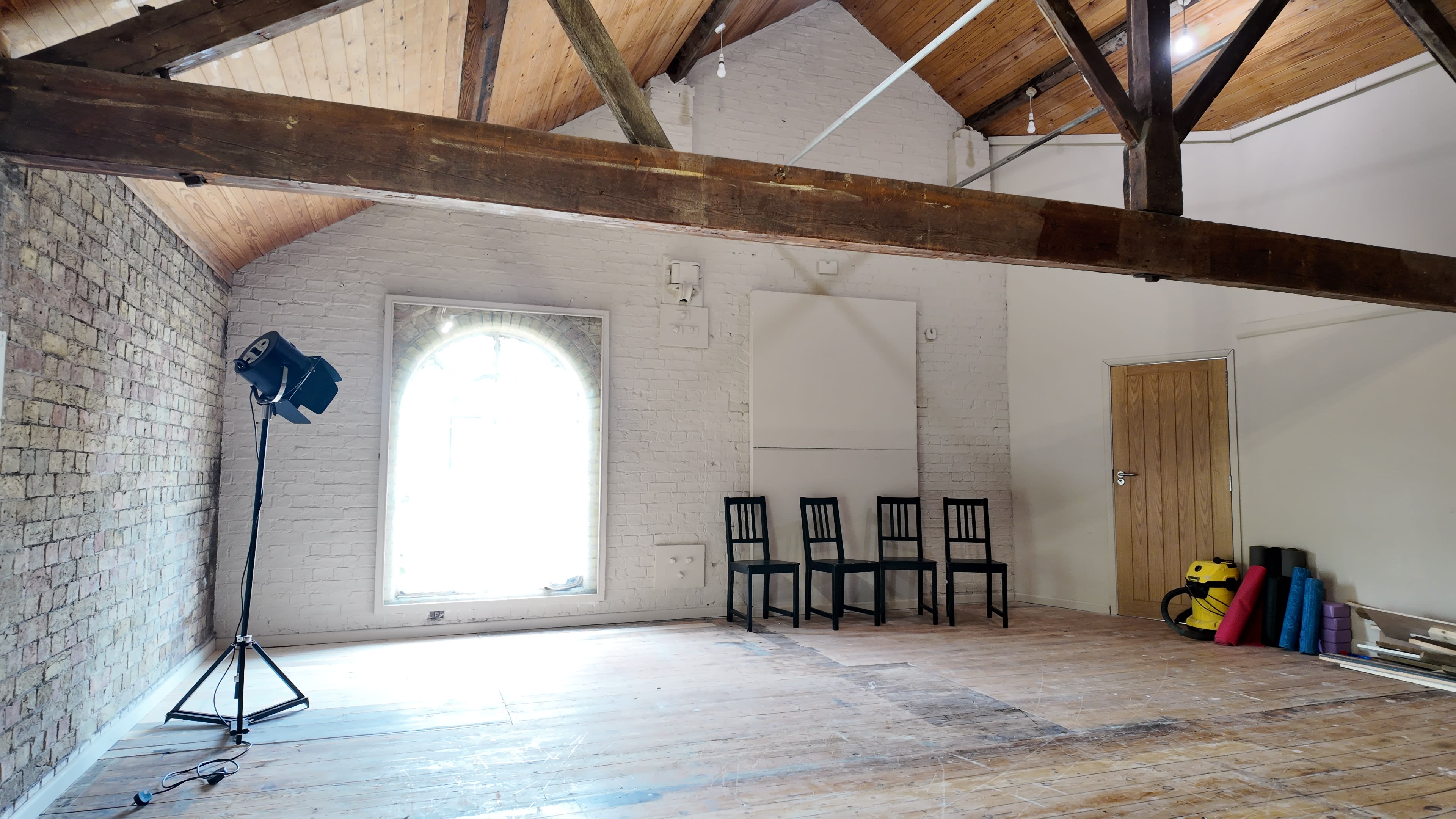 The image shows an empty room with wooden beams, a large arched window, a lighting setup on a stand, four black chairs against the wall, and various exercise mats rolled up in a corner.