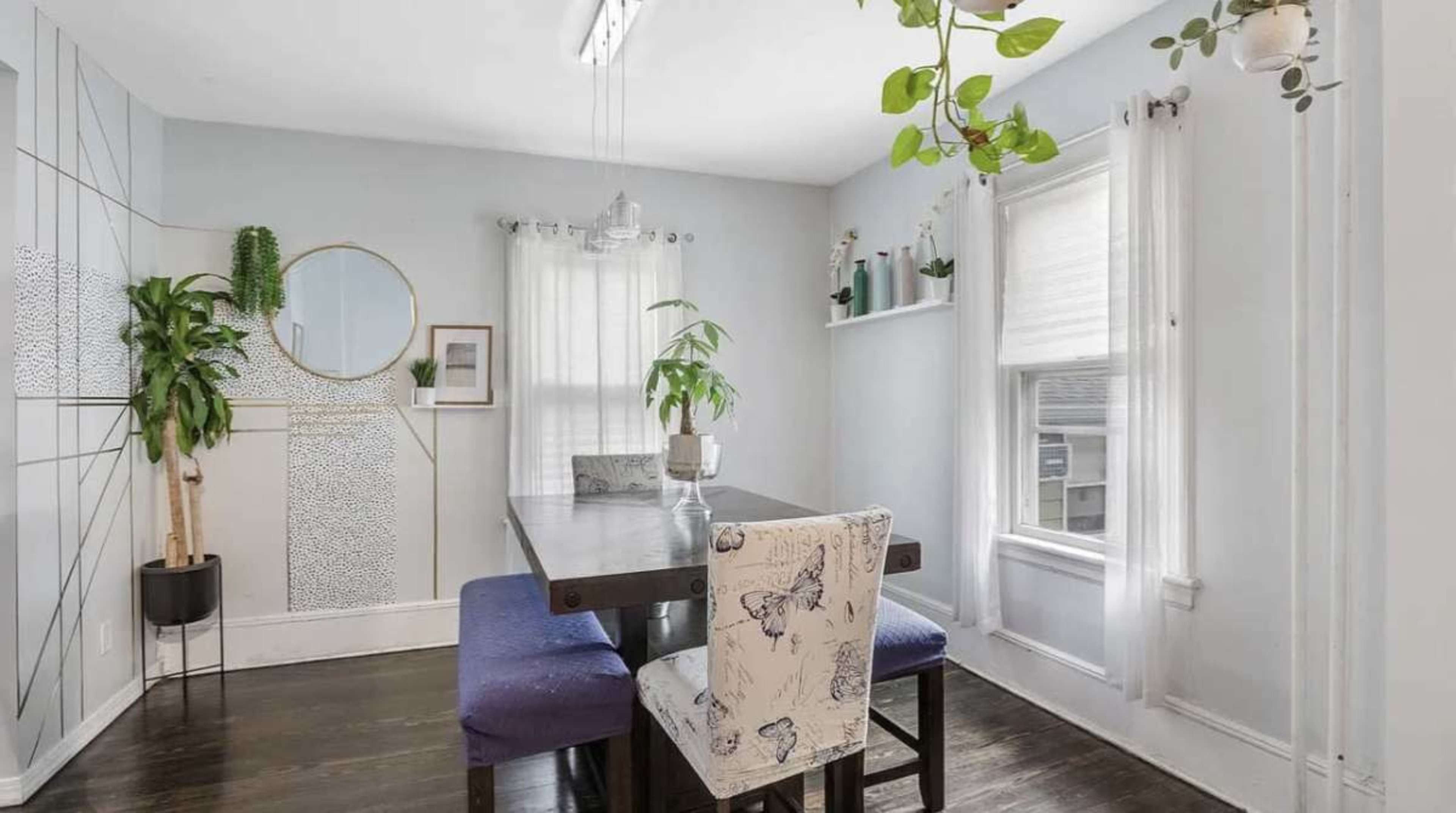 The image shows a dining area featuring a rectangular table with upholstered chairs, a large round mirror on the wall, plants near the windows, and light-colored walls.