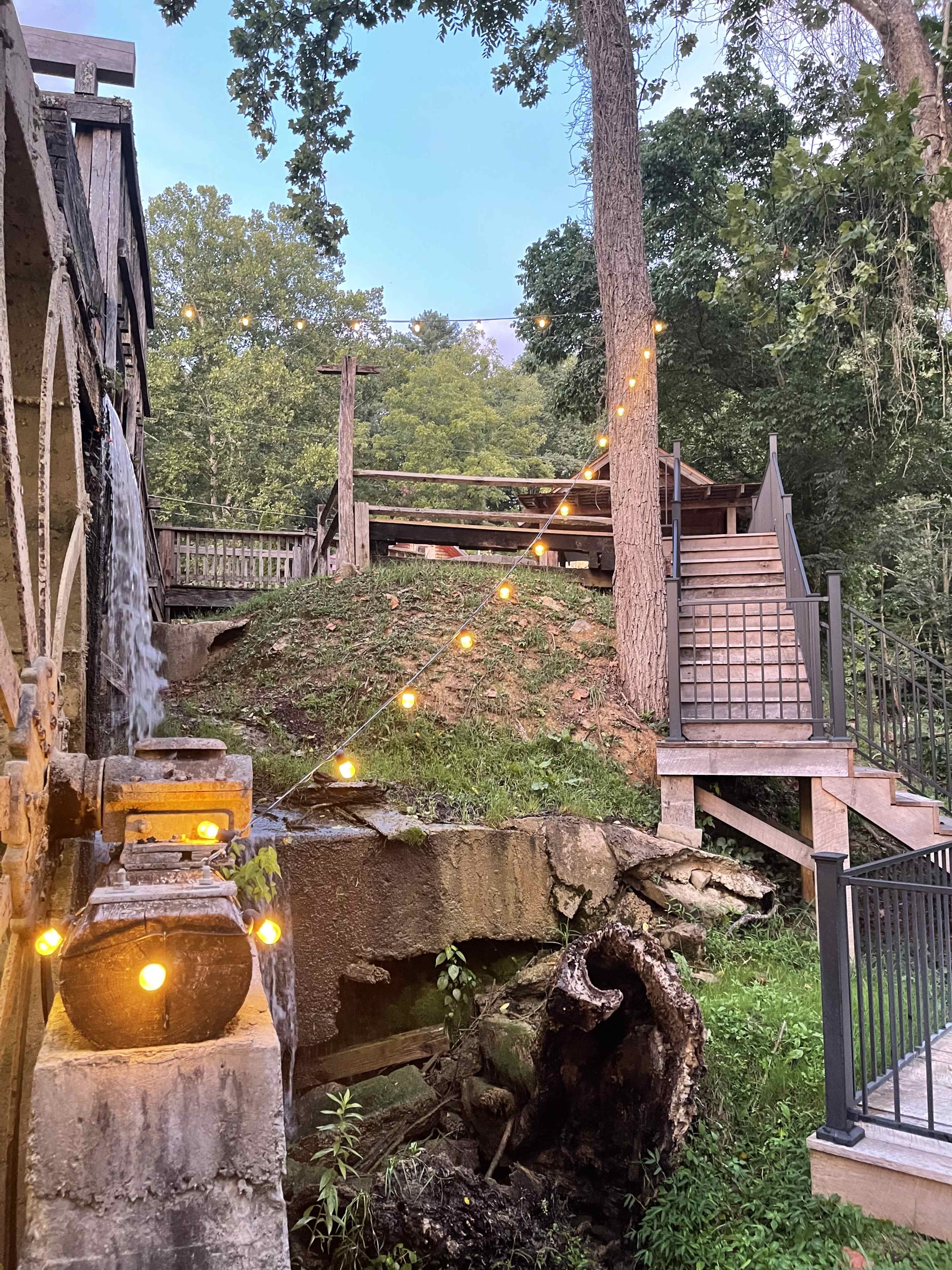 A wooden structure with a water feature and string lights is situated in a wooded area, featuring stairs that lead to a higher platform.