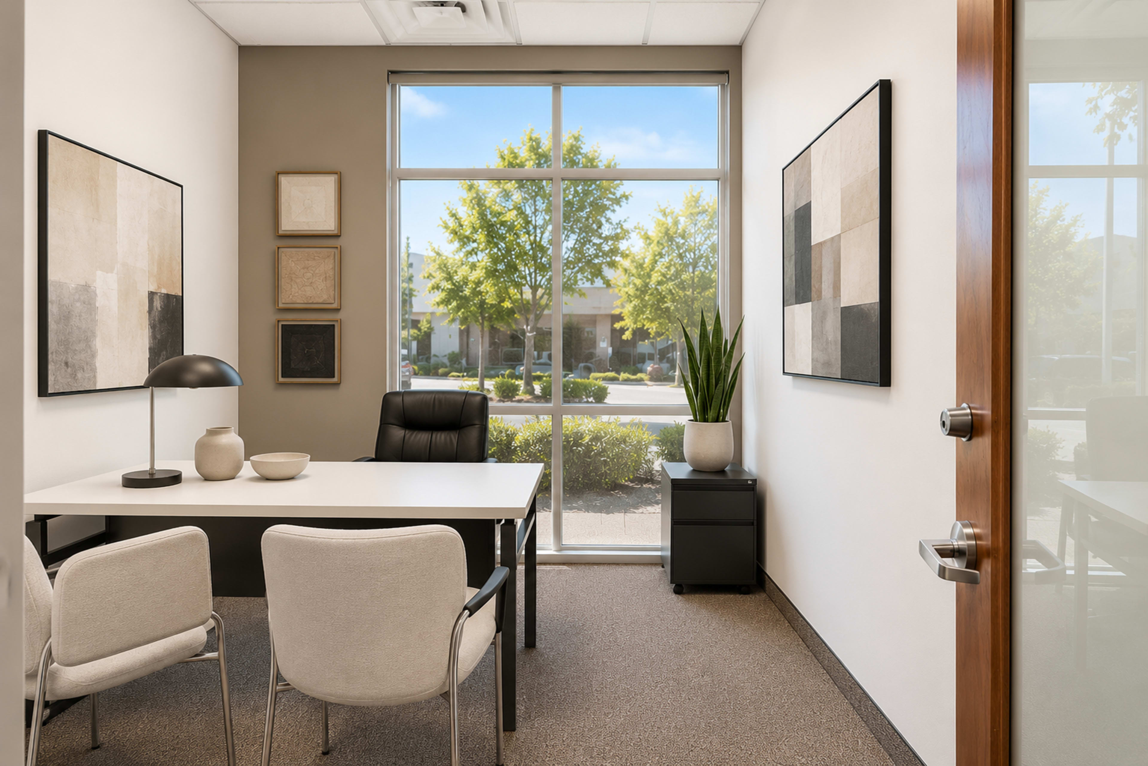The image shows a modern office space with a white desk, black chair, and two light-colored chairs, featuring large windows that provide a view of trees outside.