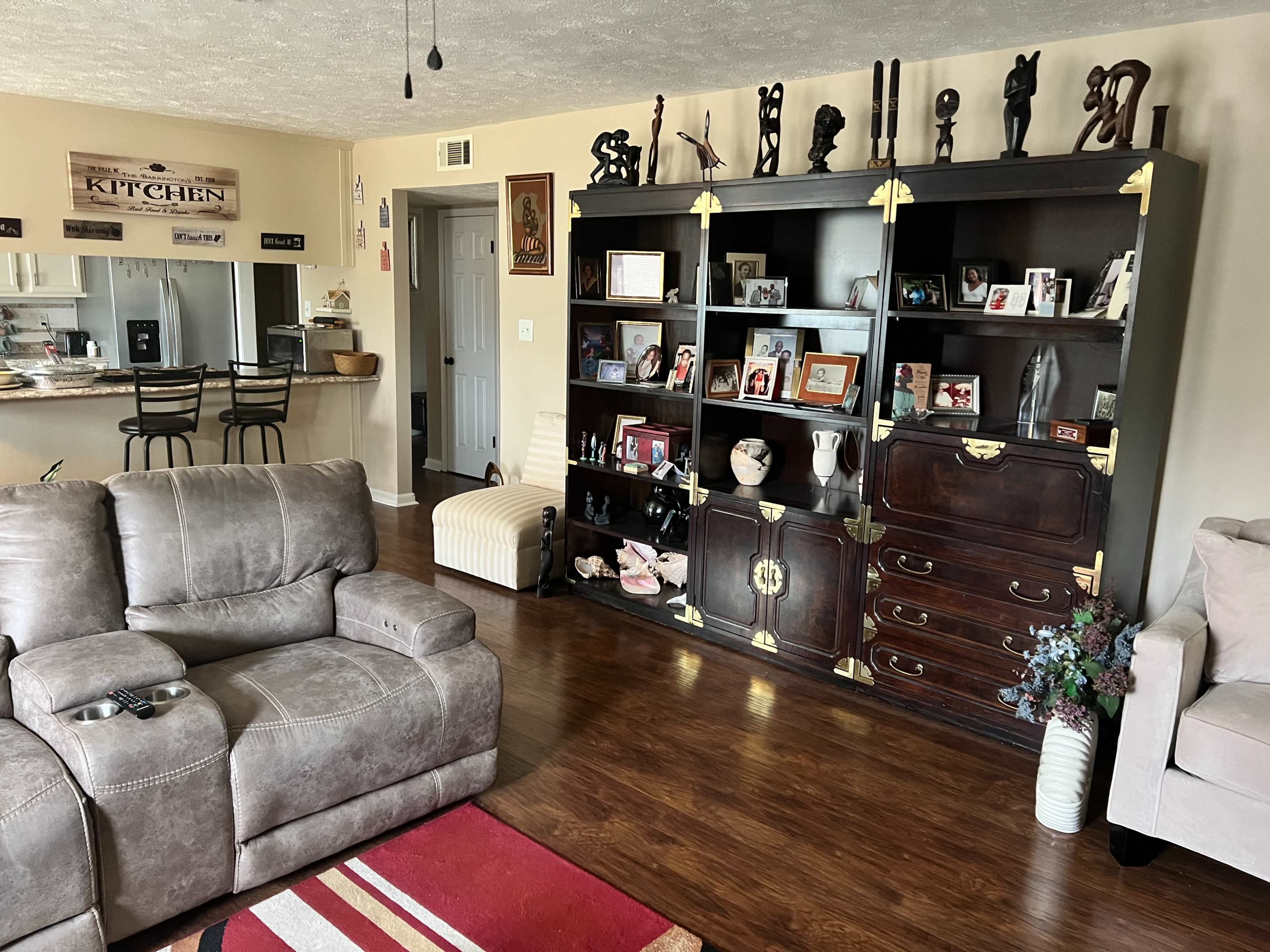 A living room with a gray sectional sofa, a wooden bookshelf filled with family photographs, and a kitchenette in the background.