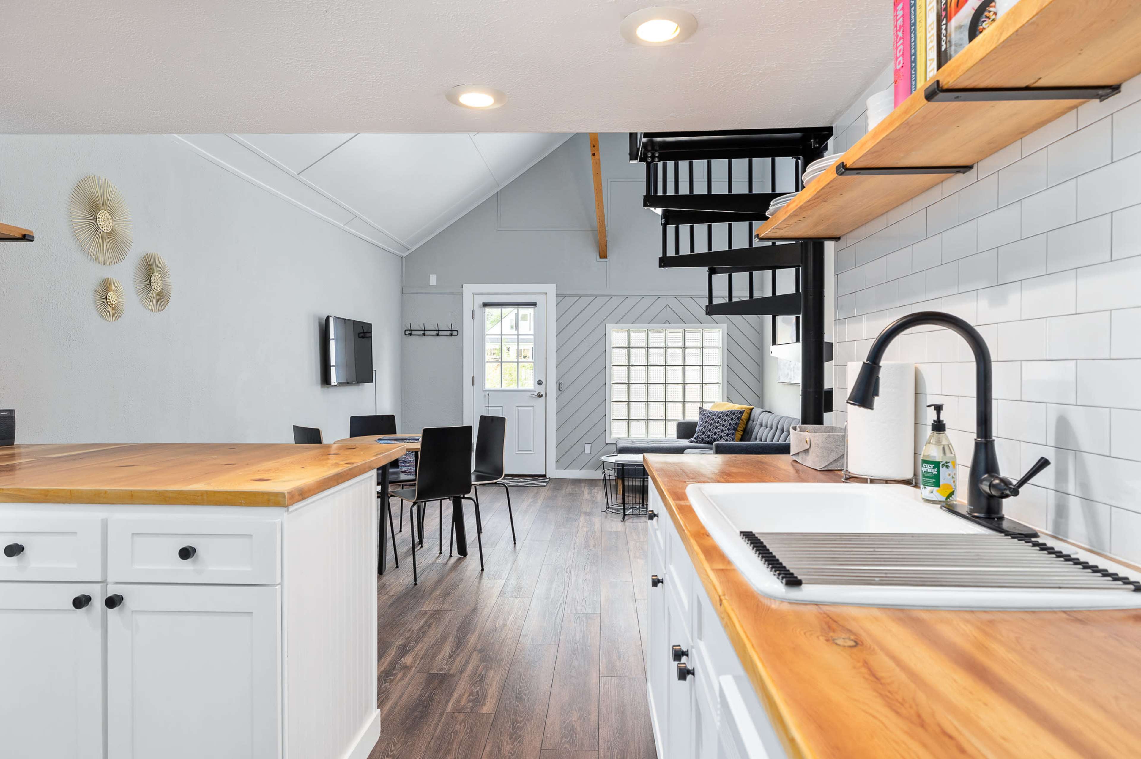 The image shows a modern kitchen with a wooden countertop, white cabinets, and an open layout that leads to a dining area and a spiral staircase.