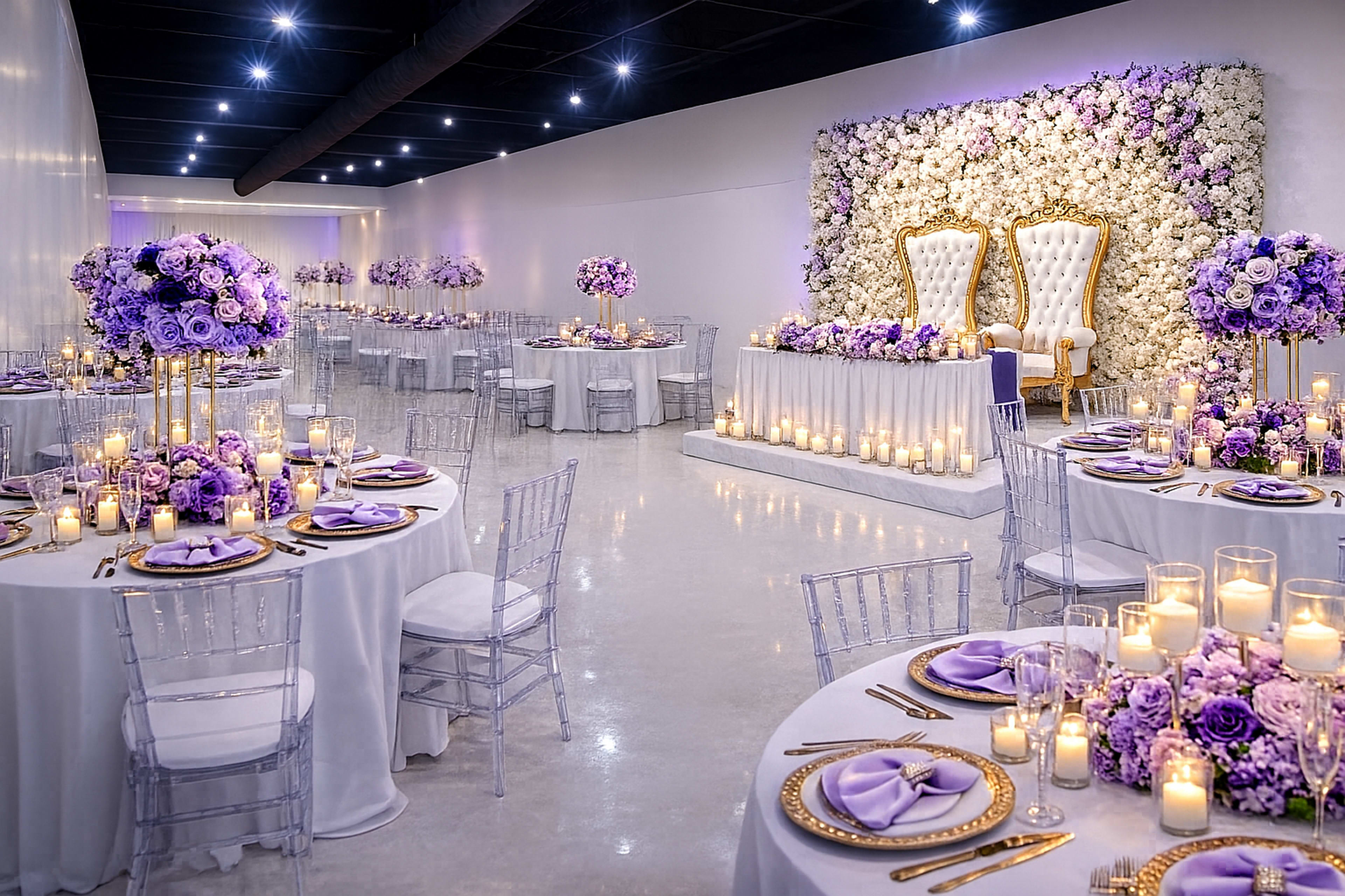 The image shows a beautifully decorated event space with round tables set with floral centerpieces, candles, and elegant tableware, featuring a floral backdrop and two ornate chairs at the front.