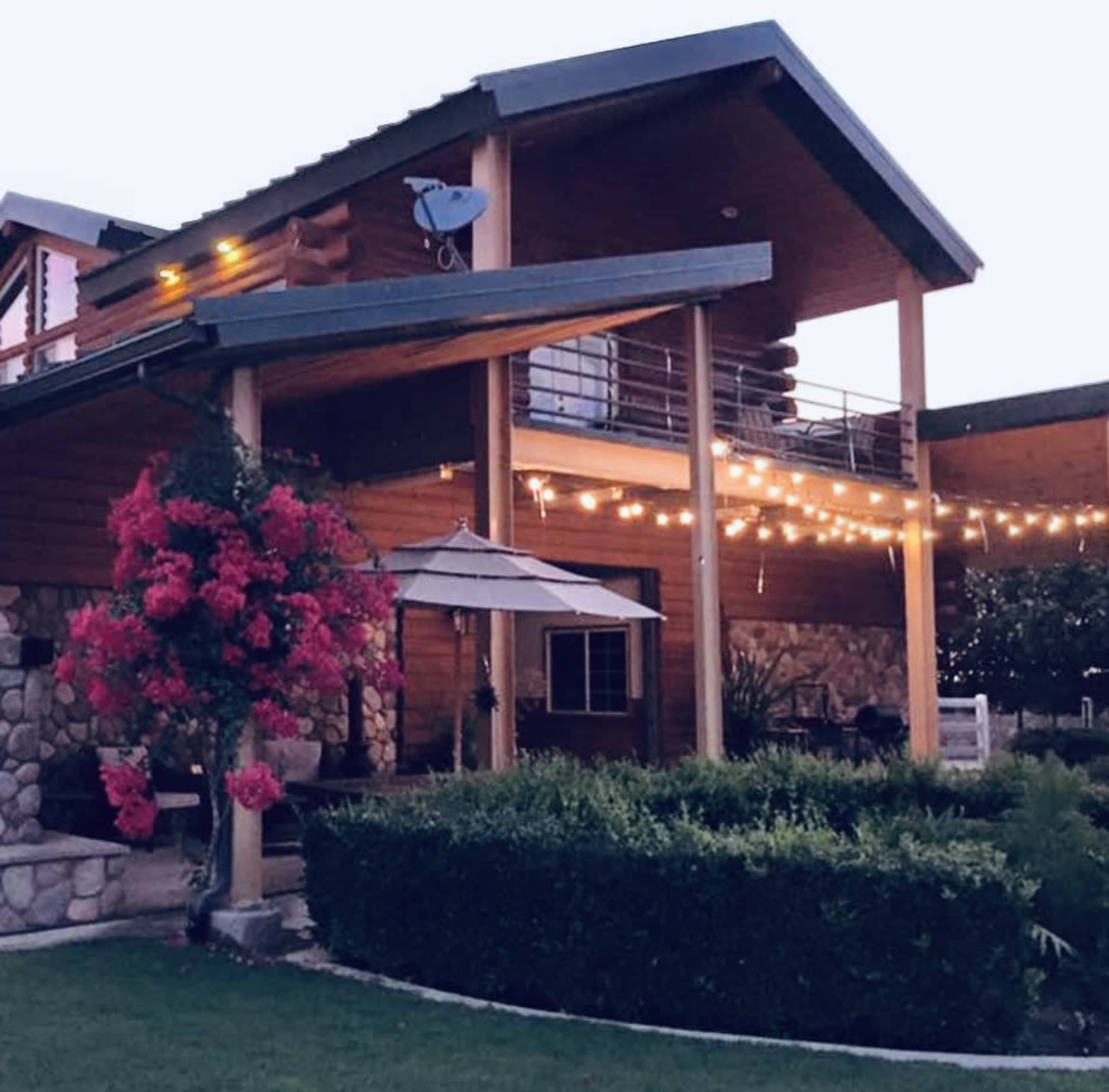 A two-story wooden house features a stone facade, outdoor seating area, and decorative string lights.