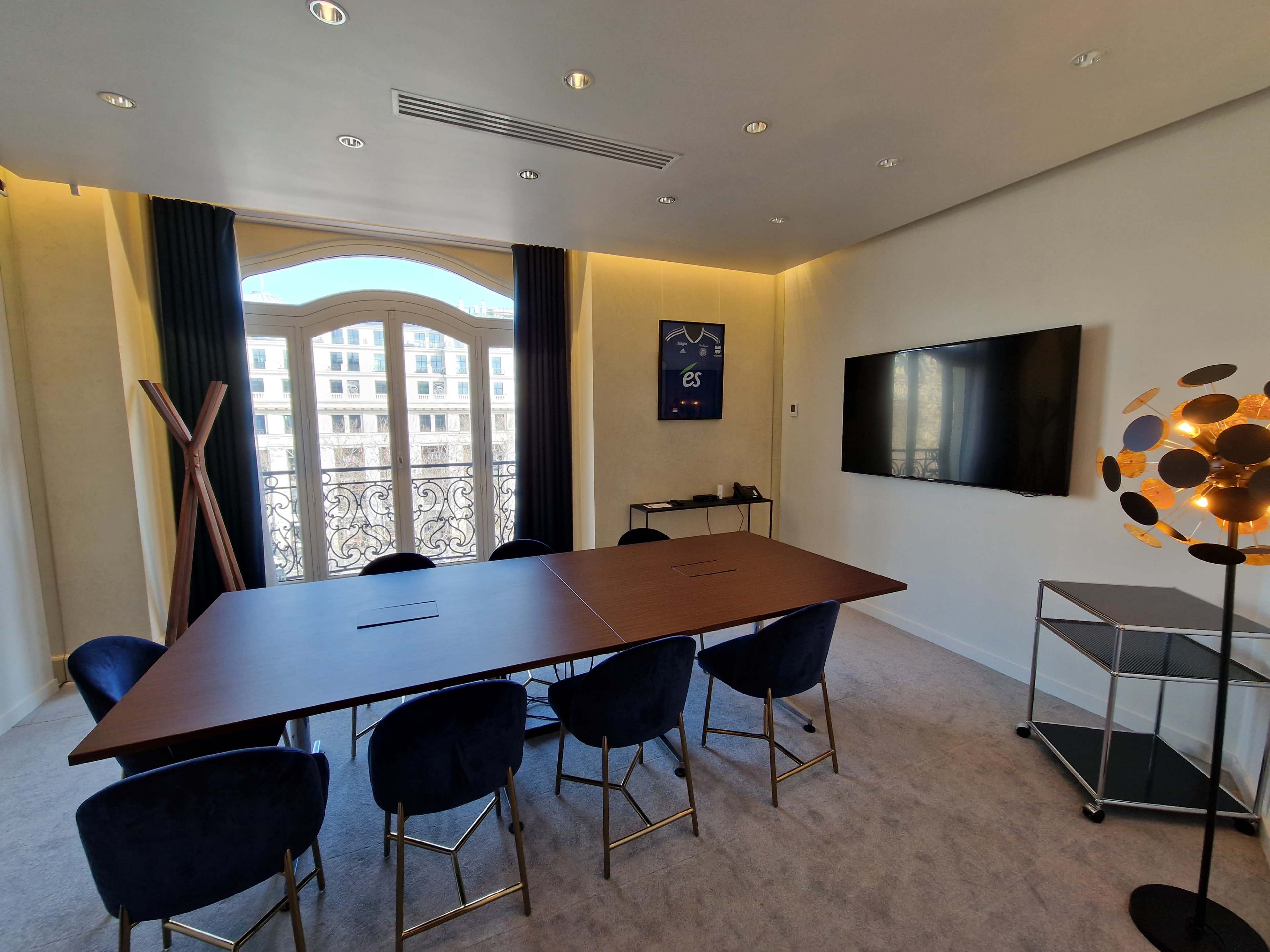 A modern conference room features a large table, plush chairs, a wall-mounted display, and a balcony with a view.