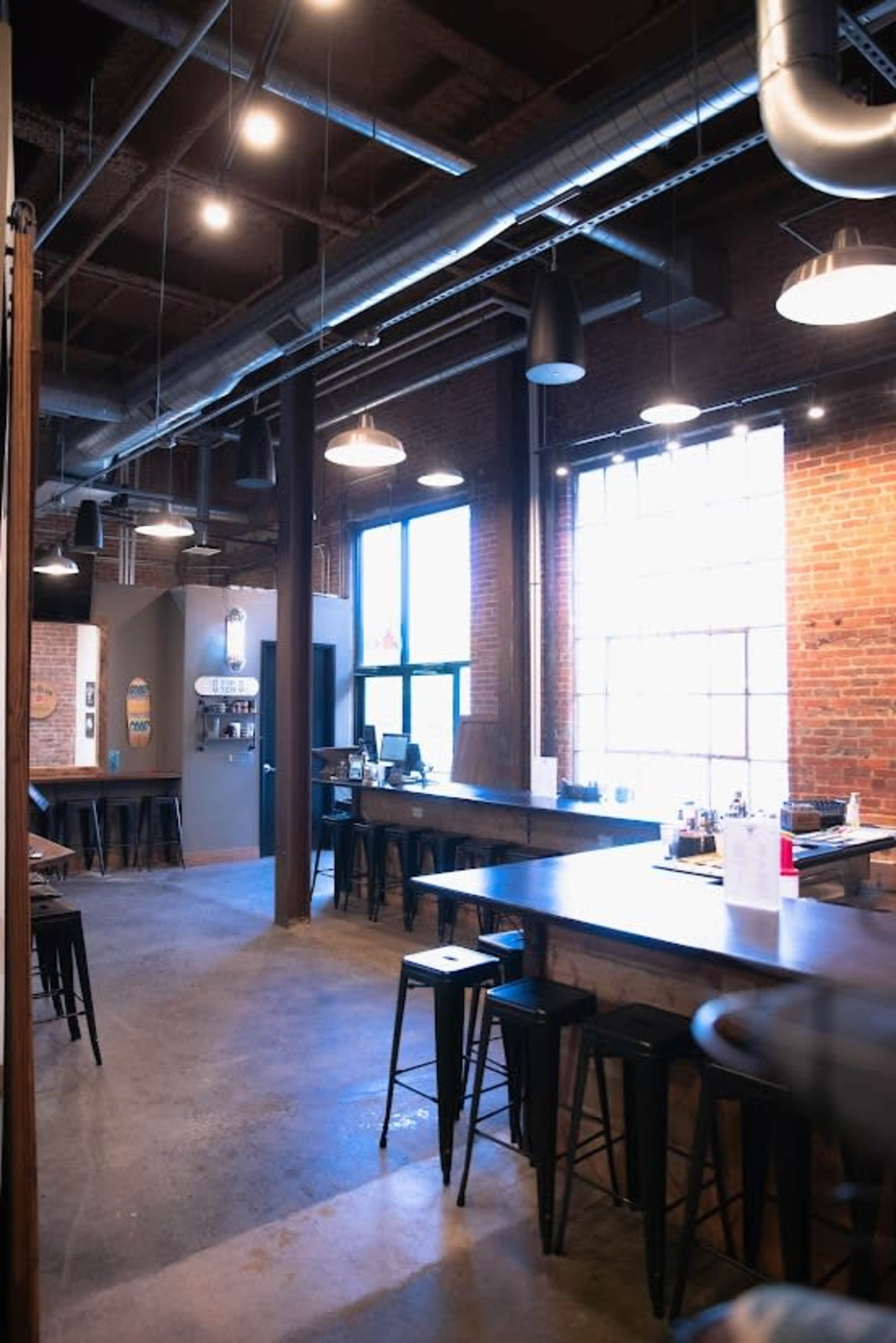 The image shows a spacious industrial-style cafe with exposed brick walls, high ceilings, and a long bar counter lined with stools.