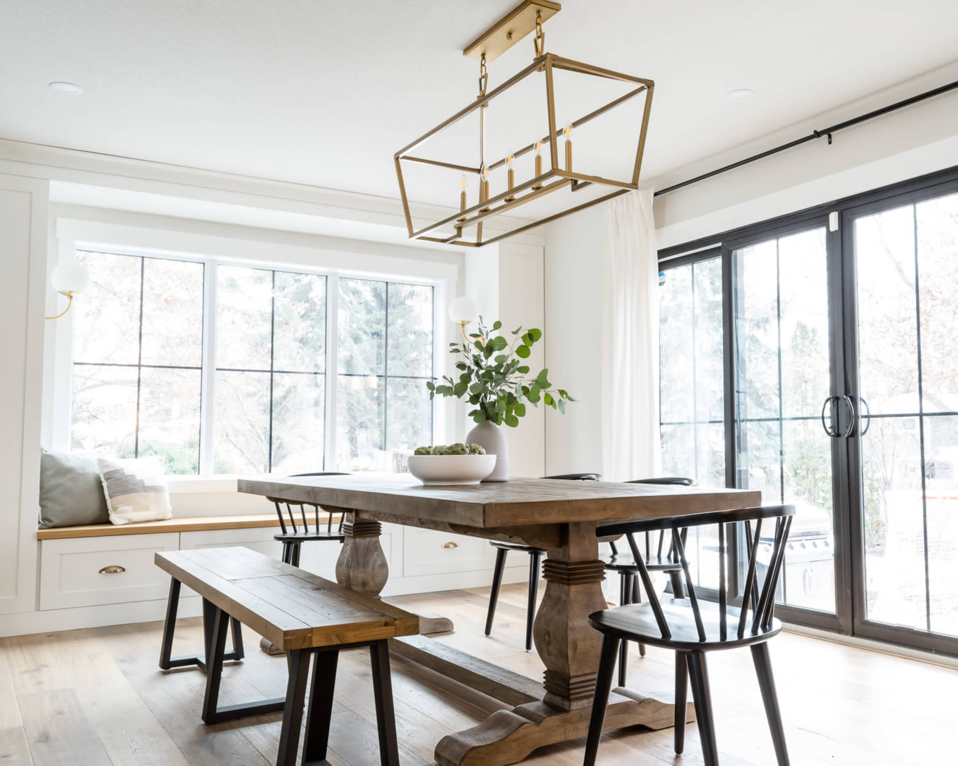 A dining area features a wooden table with a bench and chairs, illuminated by a rectangular gold light fixture, near large windows with natural light.