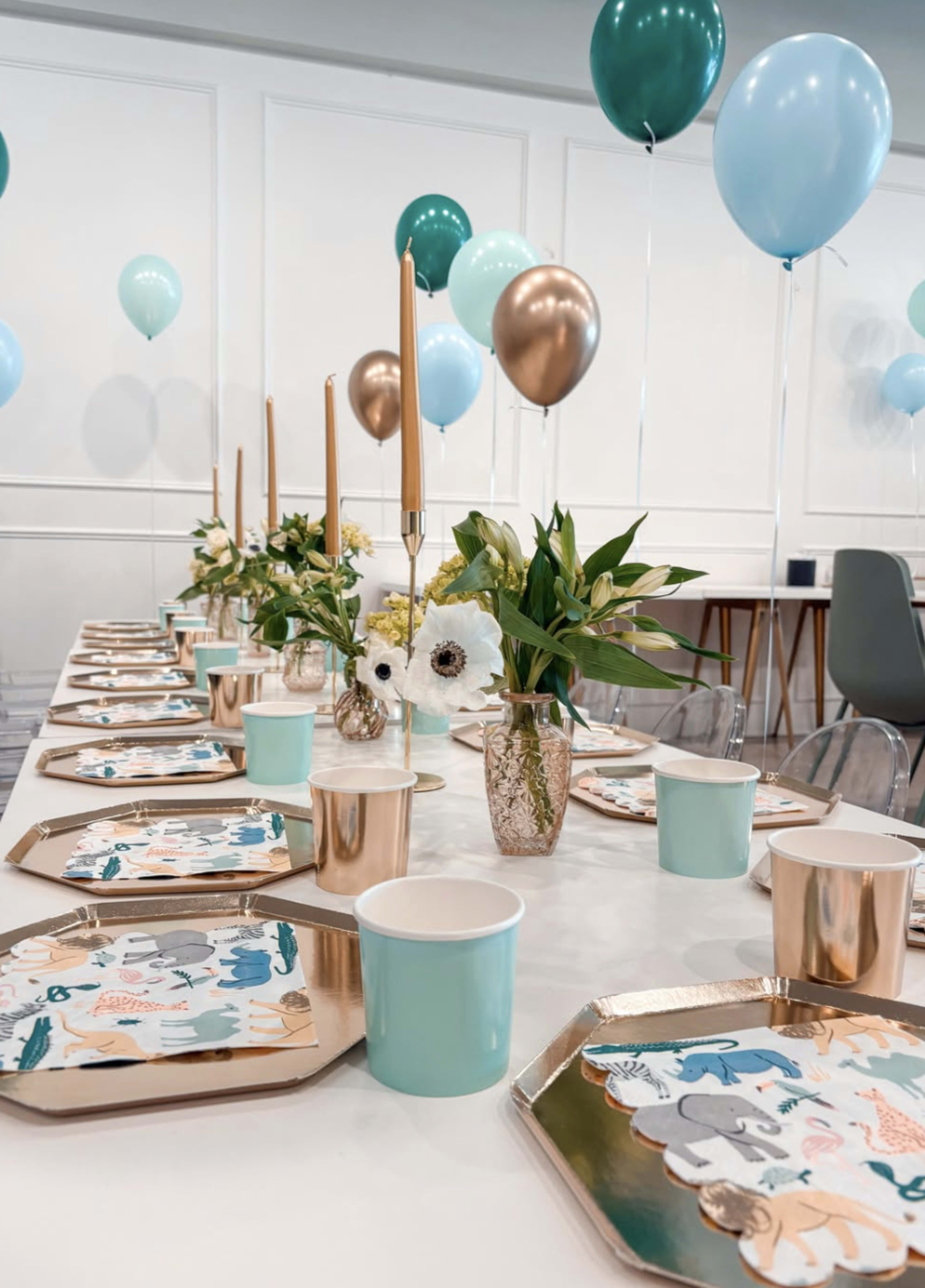 The image shows a decorated dining table set for an event, featuring pastel balloons, gold candle holders, floral arrangements, and themed tableware.