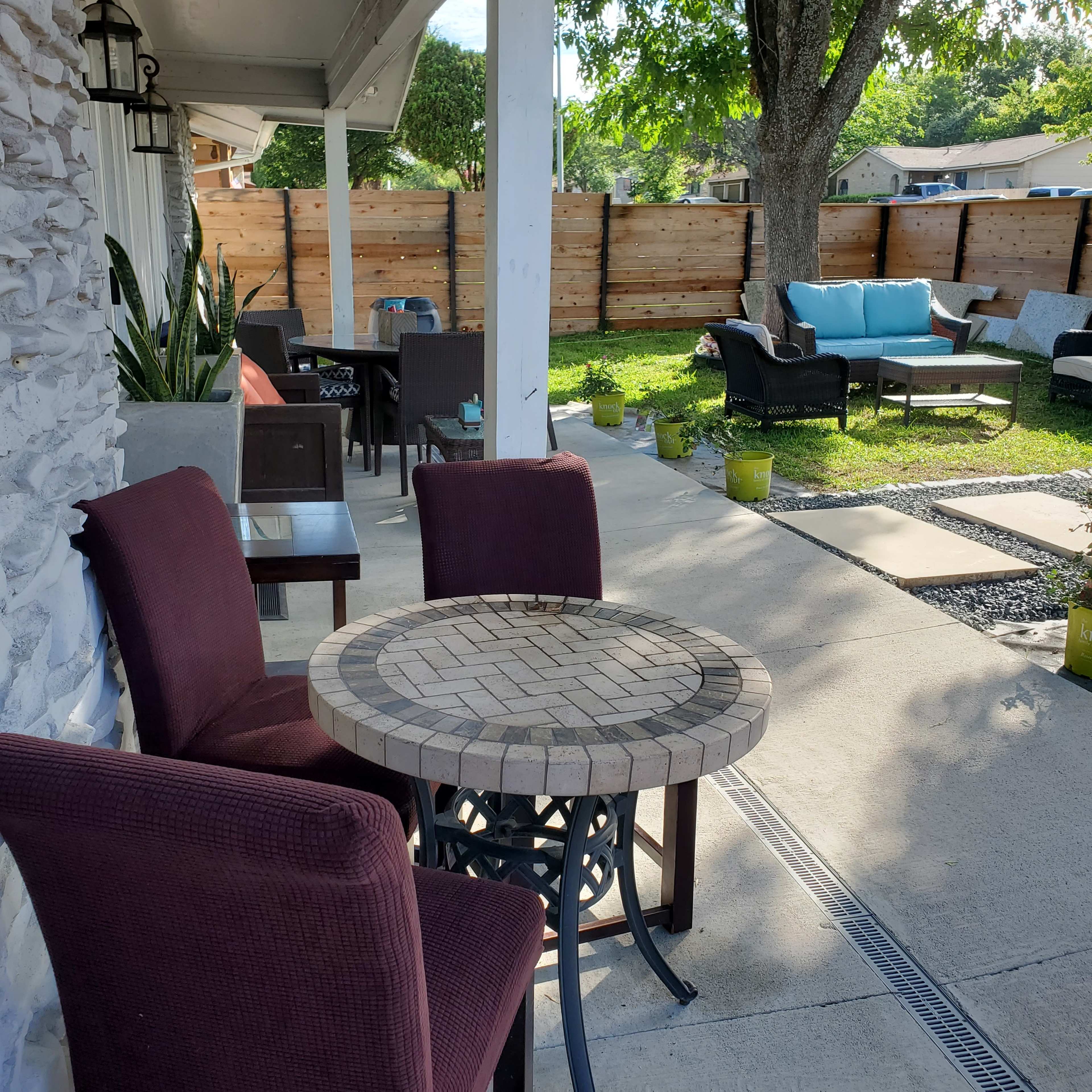 A patio area features a round stone table with two burgundy chairs, surrounded by various seating arrangements and potted plants.