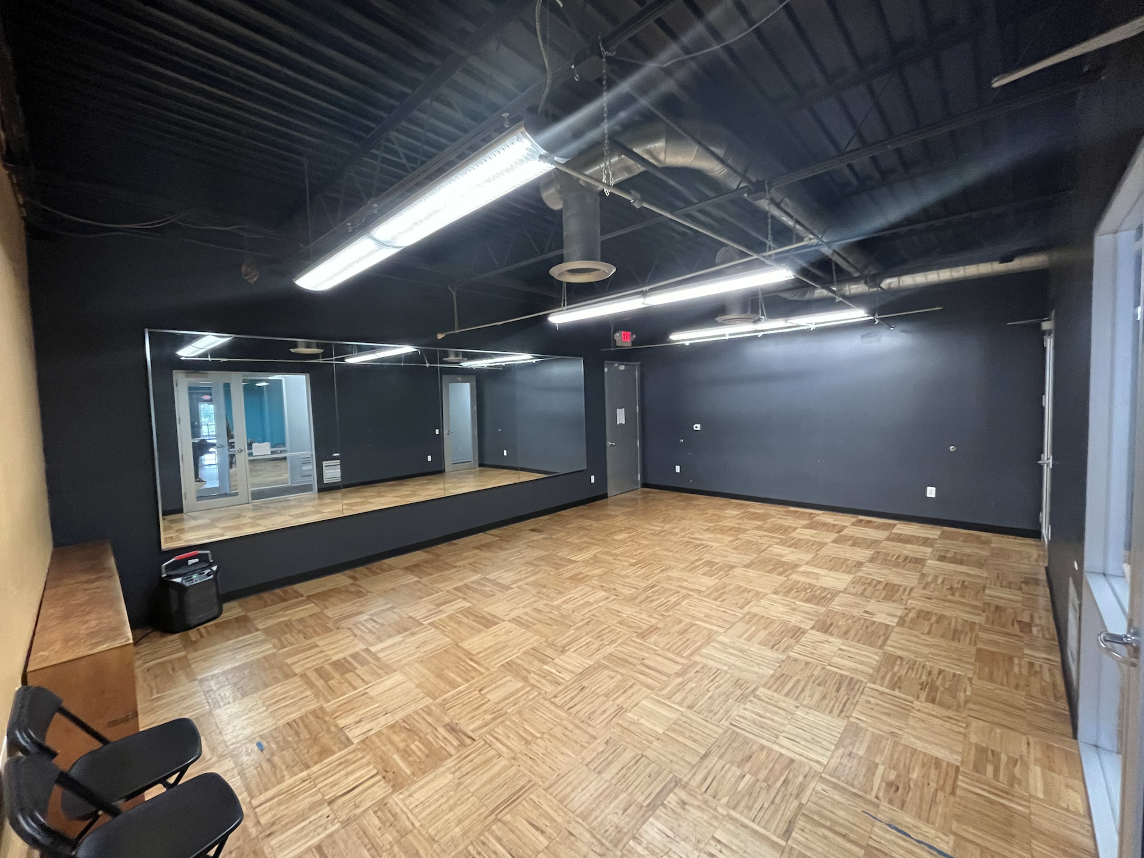 An empty studio room with dark walls, a large mirror, and wooden flooring, featuring two chairs and a trash can.