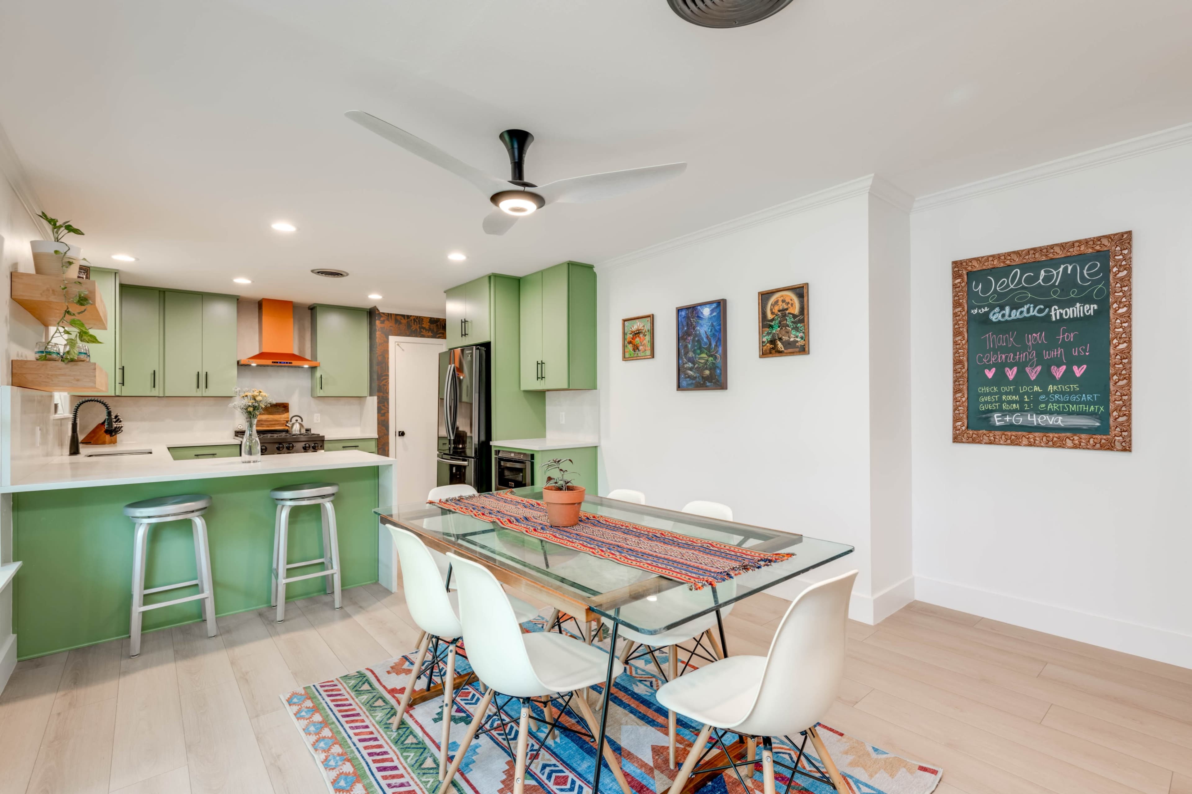 A modern kitchen features green cabinets, a glass dining table, and a colorful rug, with a chalkboard welcoming guests.
