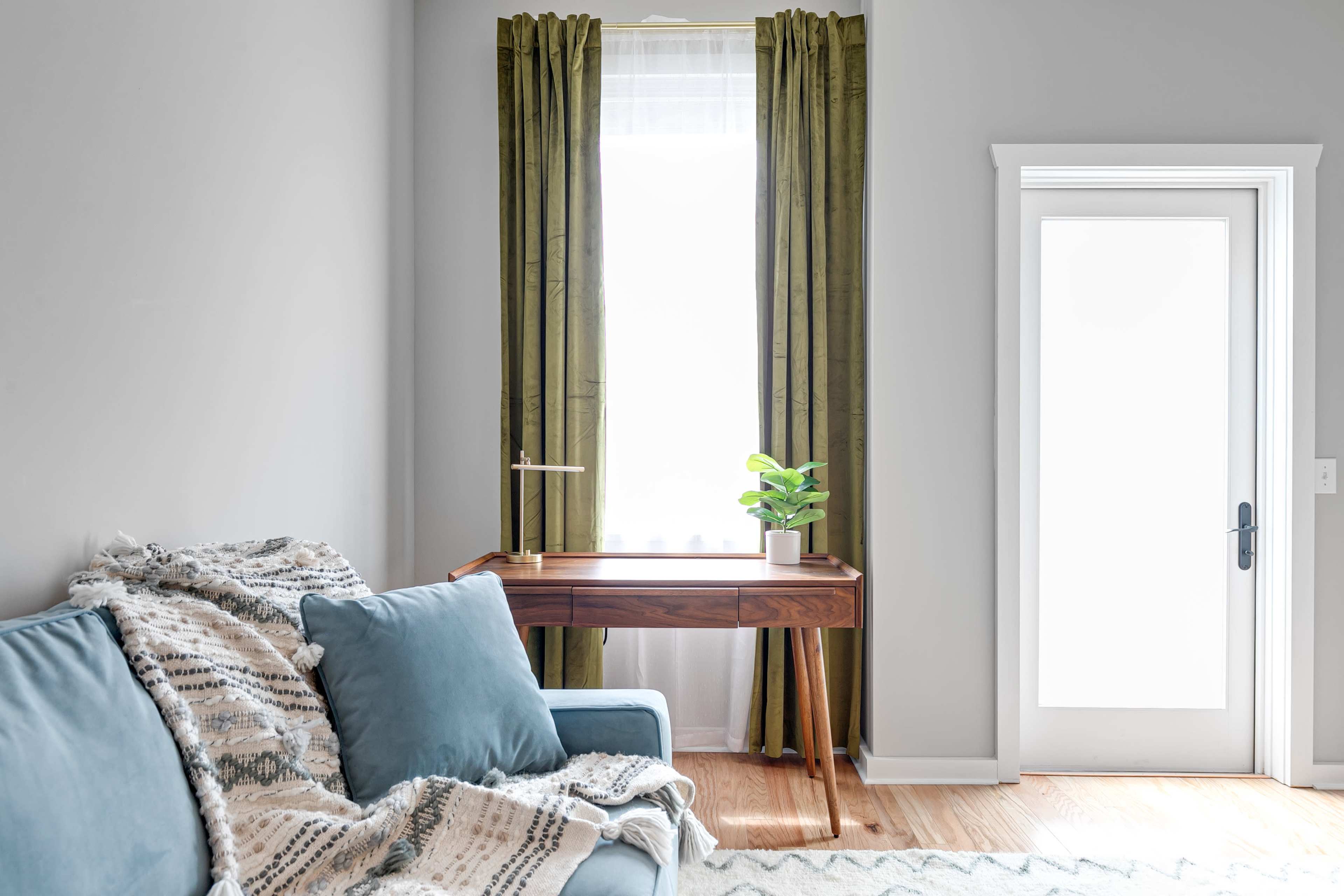 A cozy living room features a blue sofa with a patterned throw, a wooden desk with a plant, and green curtains by a window.