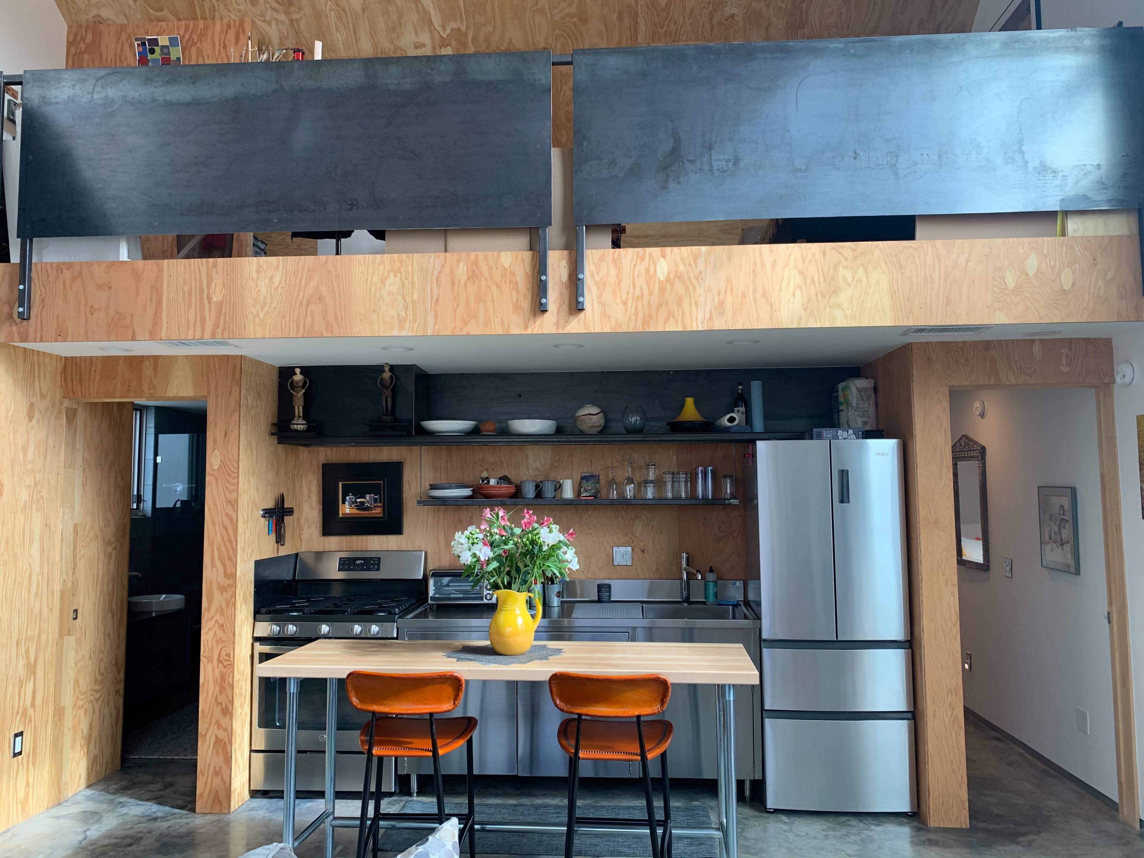 A modern kitchen with a central island, orange bar stools, and a balcony above, featuring wooden walls and stainless steel appliances.