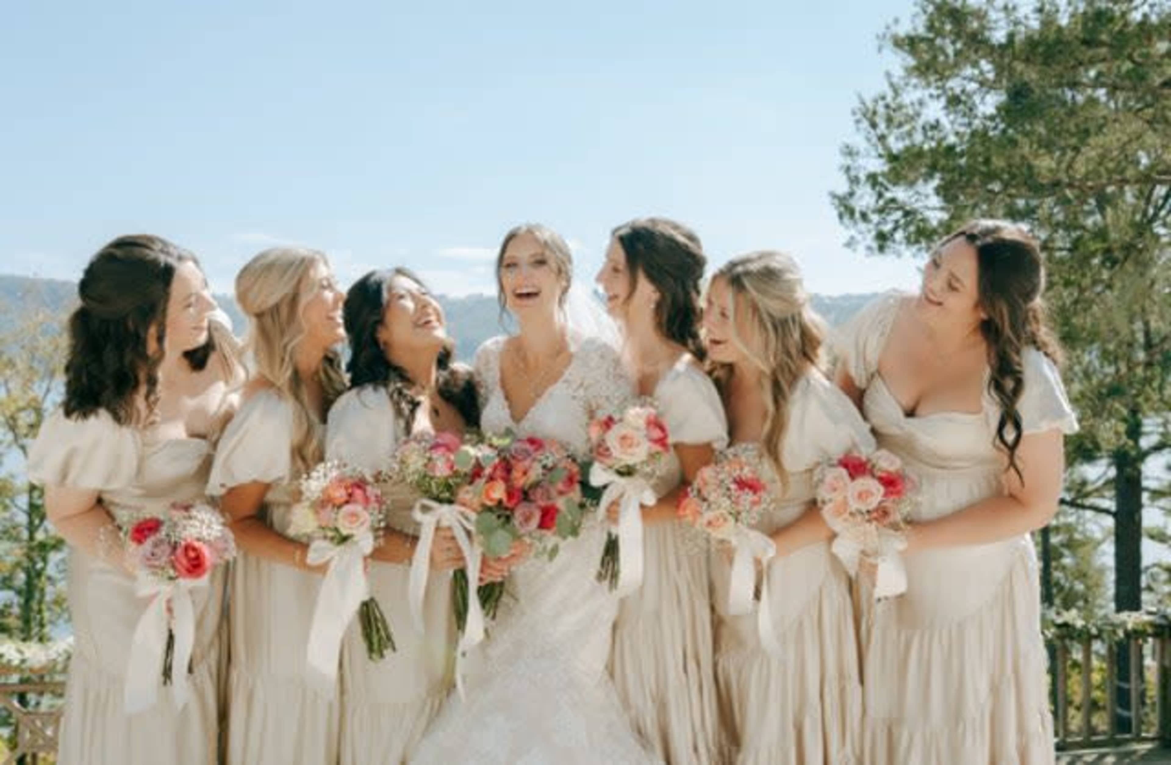 A bride stands in the center surrounded by seven bridesmaids, all holding bouquets and smiling against a backdrop of trees and a lake.