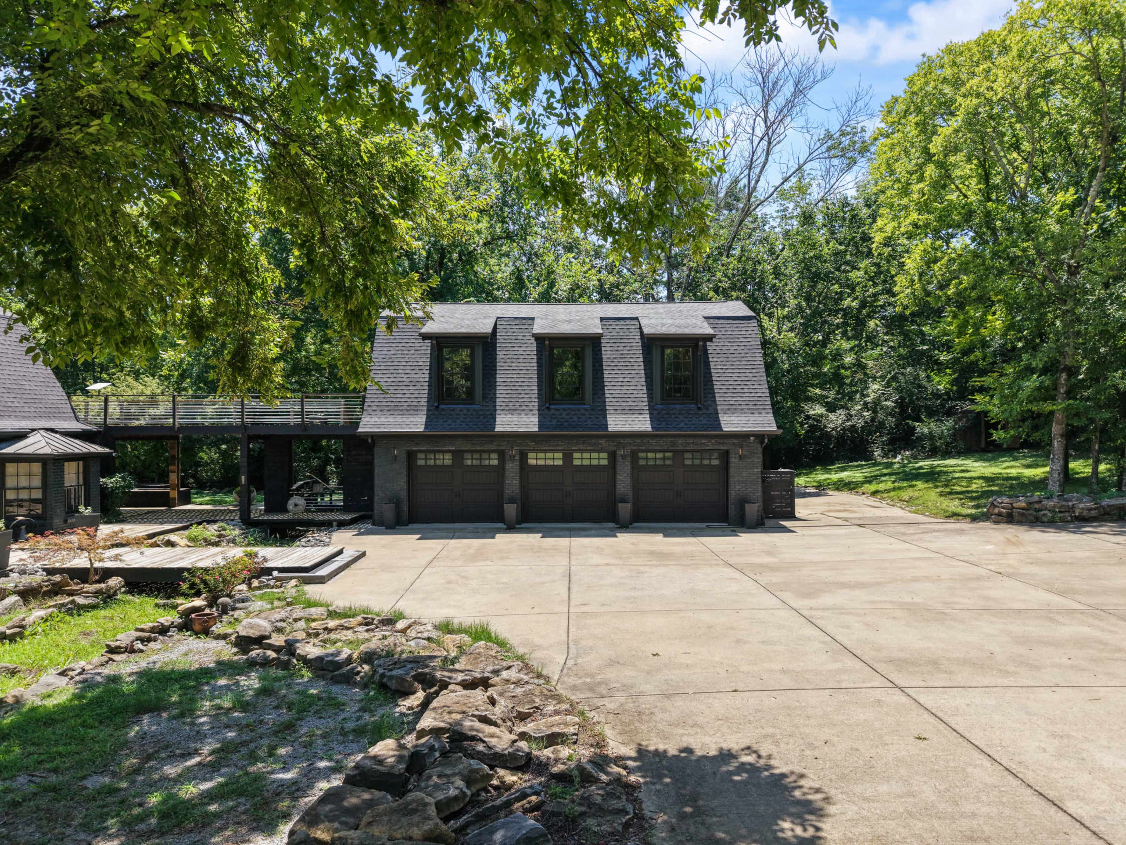 The image shows a large, two-story house with a three-car garage, surrounded by trees and a concrete driveway.
