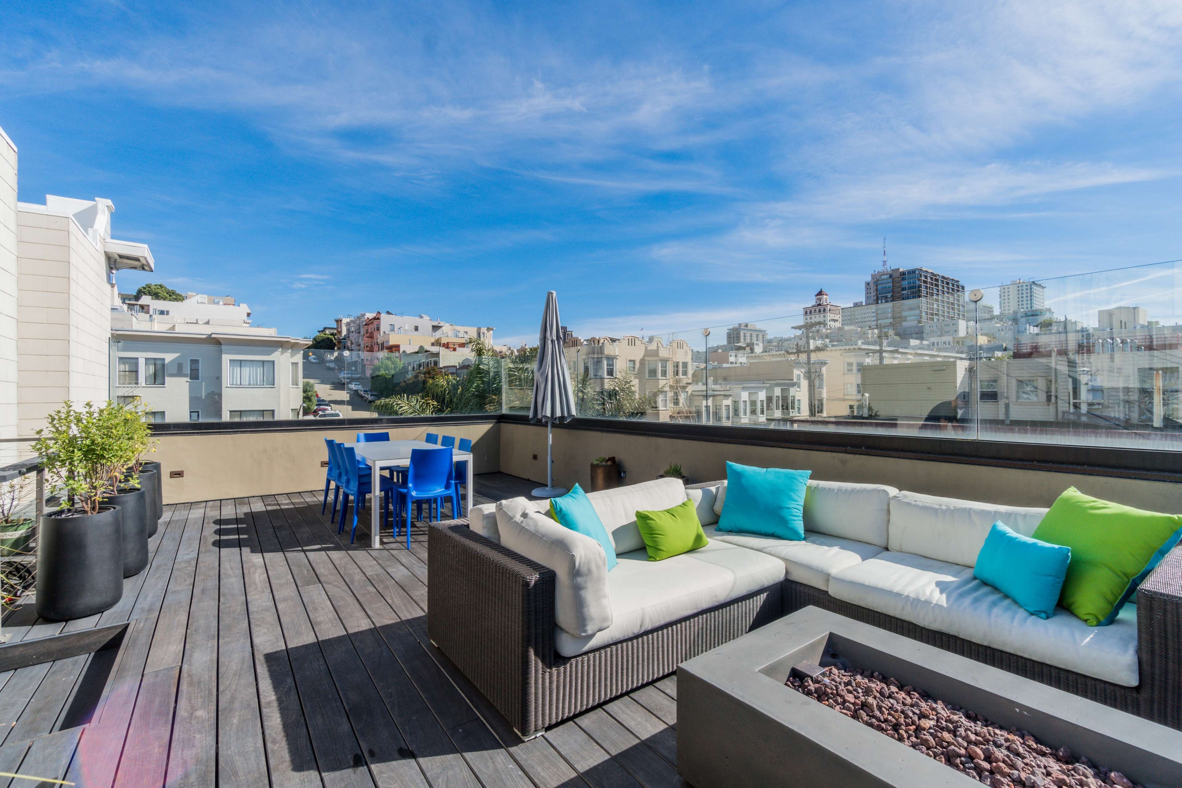 A rooftop terrace featuring a sectional sofa with colorful cushions, a dining table with blue chairs, and a clear view of city buildings under a blue sky.
