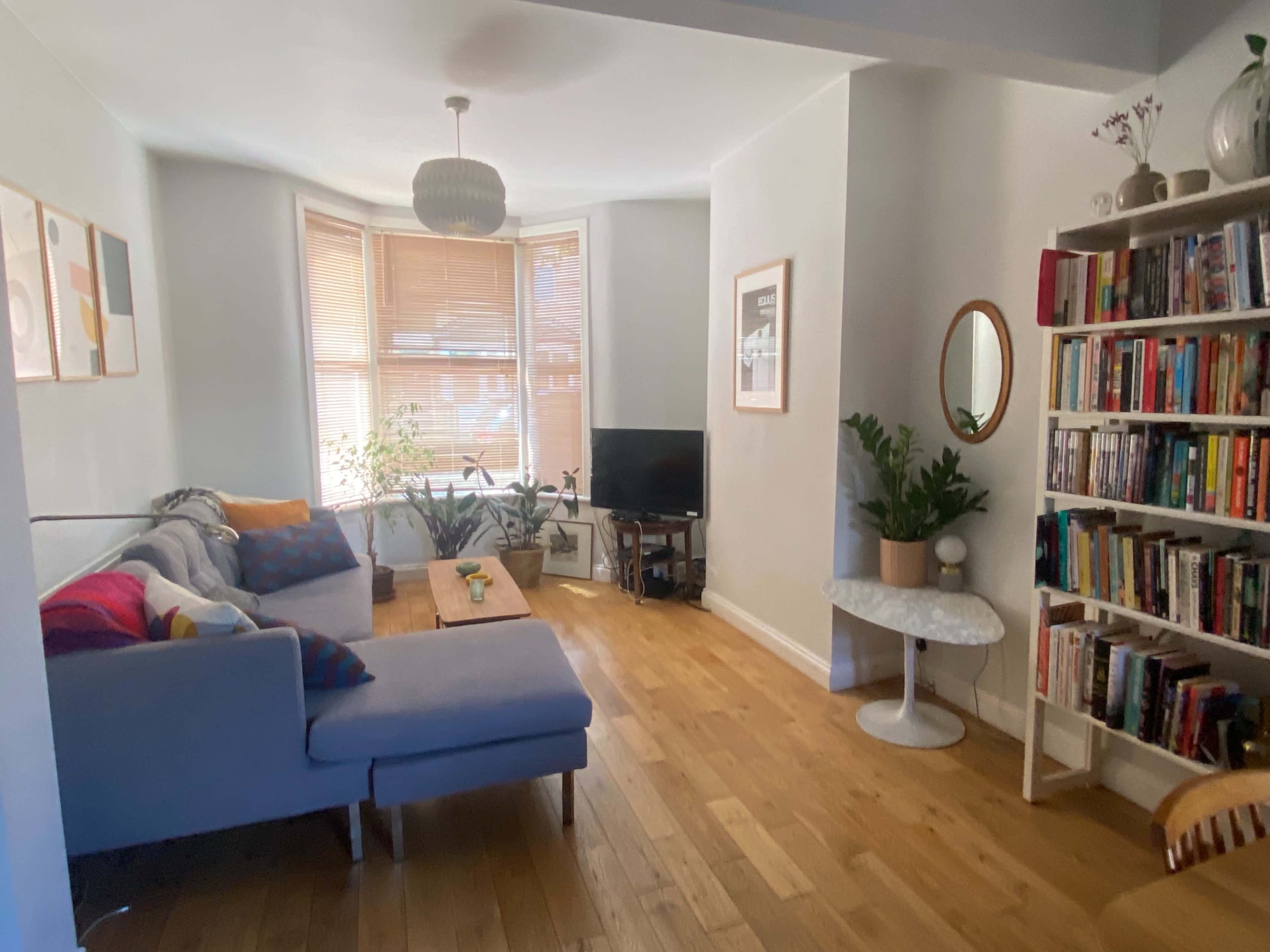 A cozy living room with a gray sectional sofa, a wooden coffee table, a television, and a bookshelf filled with books, along with plants by the window.