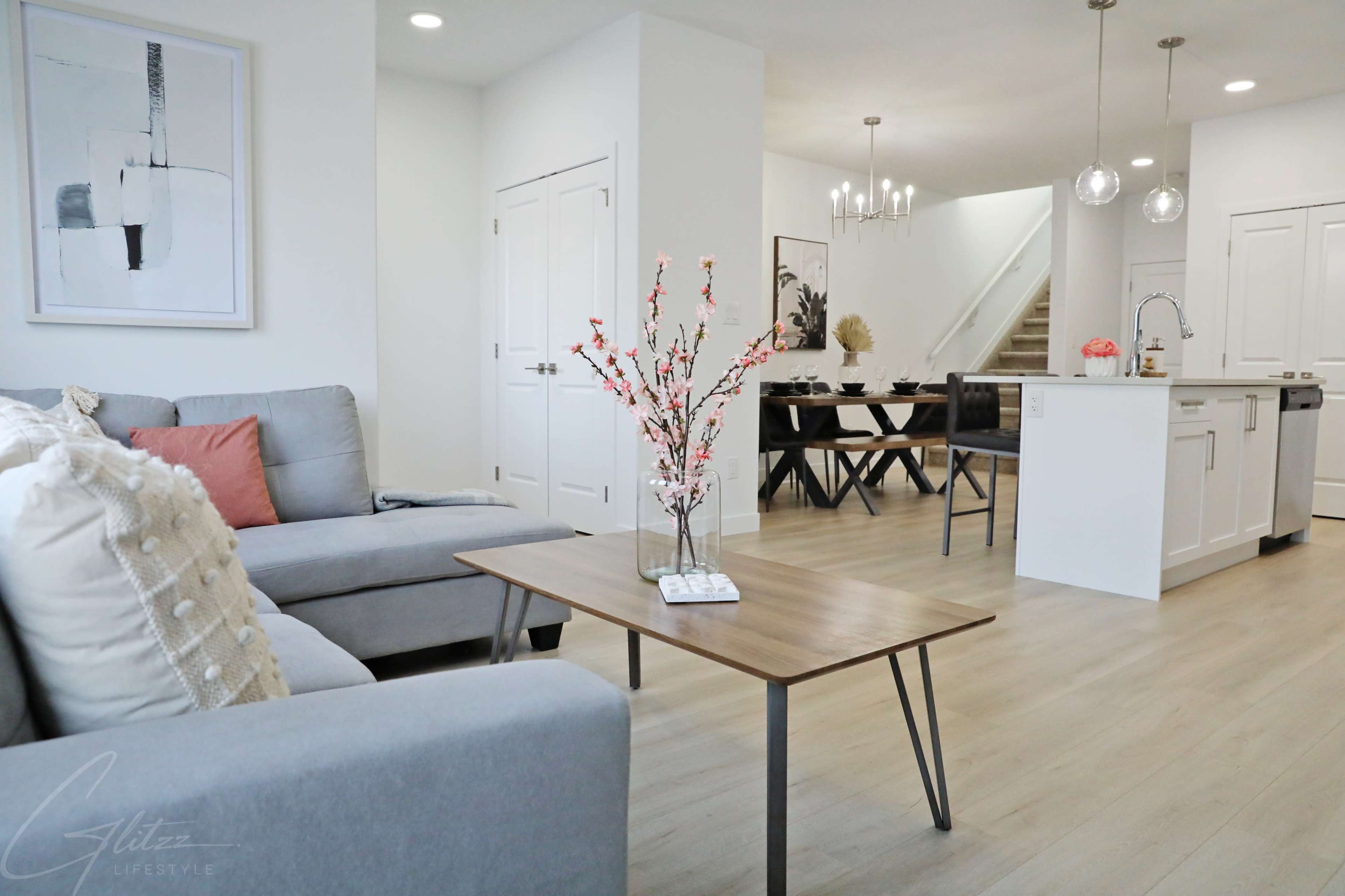 A modern living room with a gray sectional sofa, a wooden coffee table with flower decor, and an open layout leading to a dining area and kitchen.