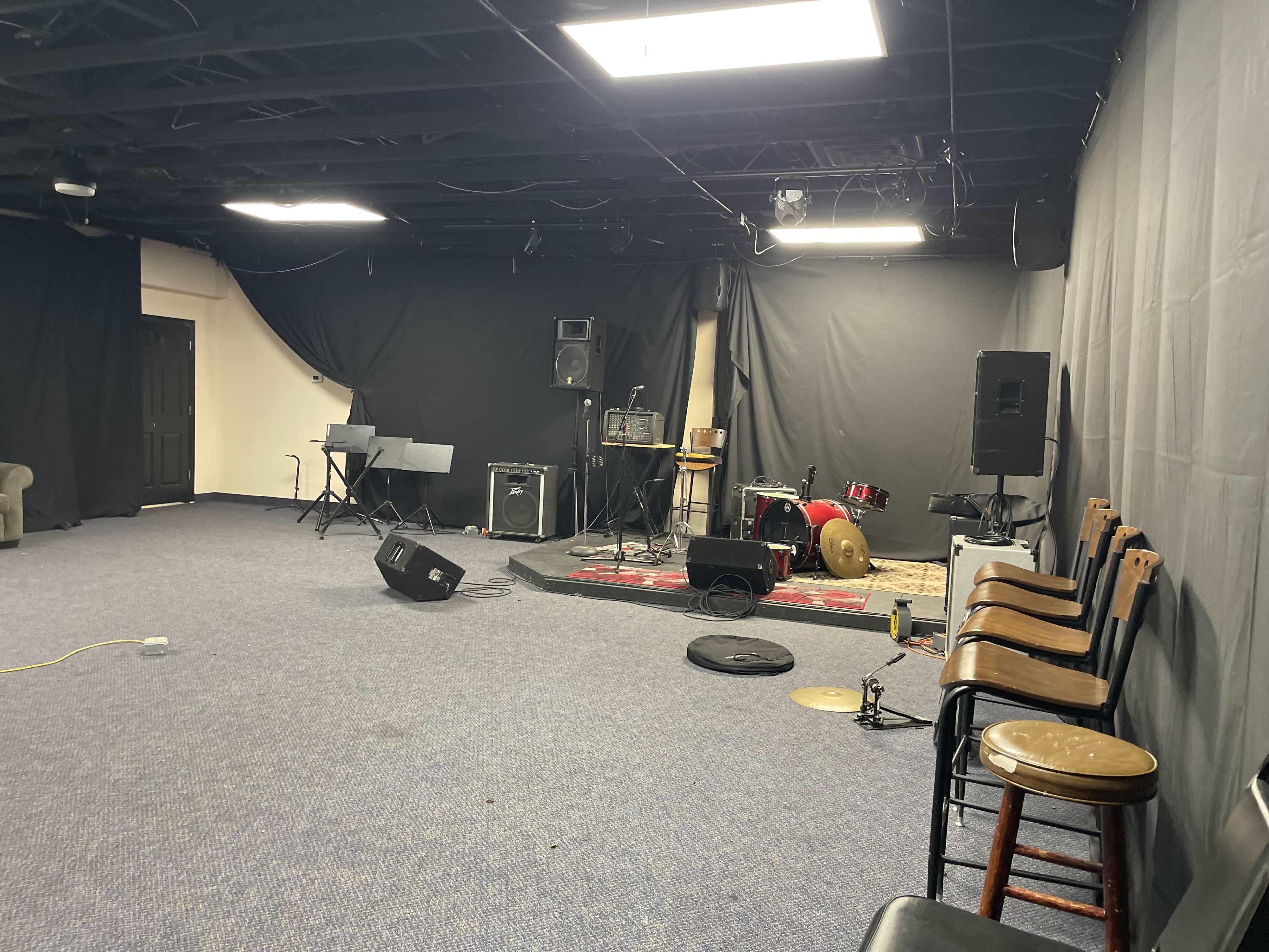 The image shows a sparse music rehearsal room with instruments, including a drum set and amplifiers, set against black curtains and a carpeted floor.