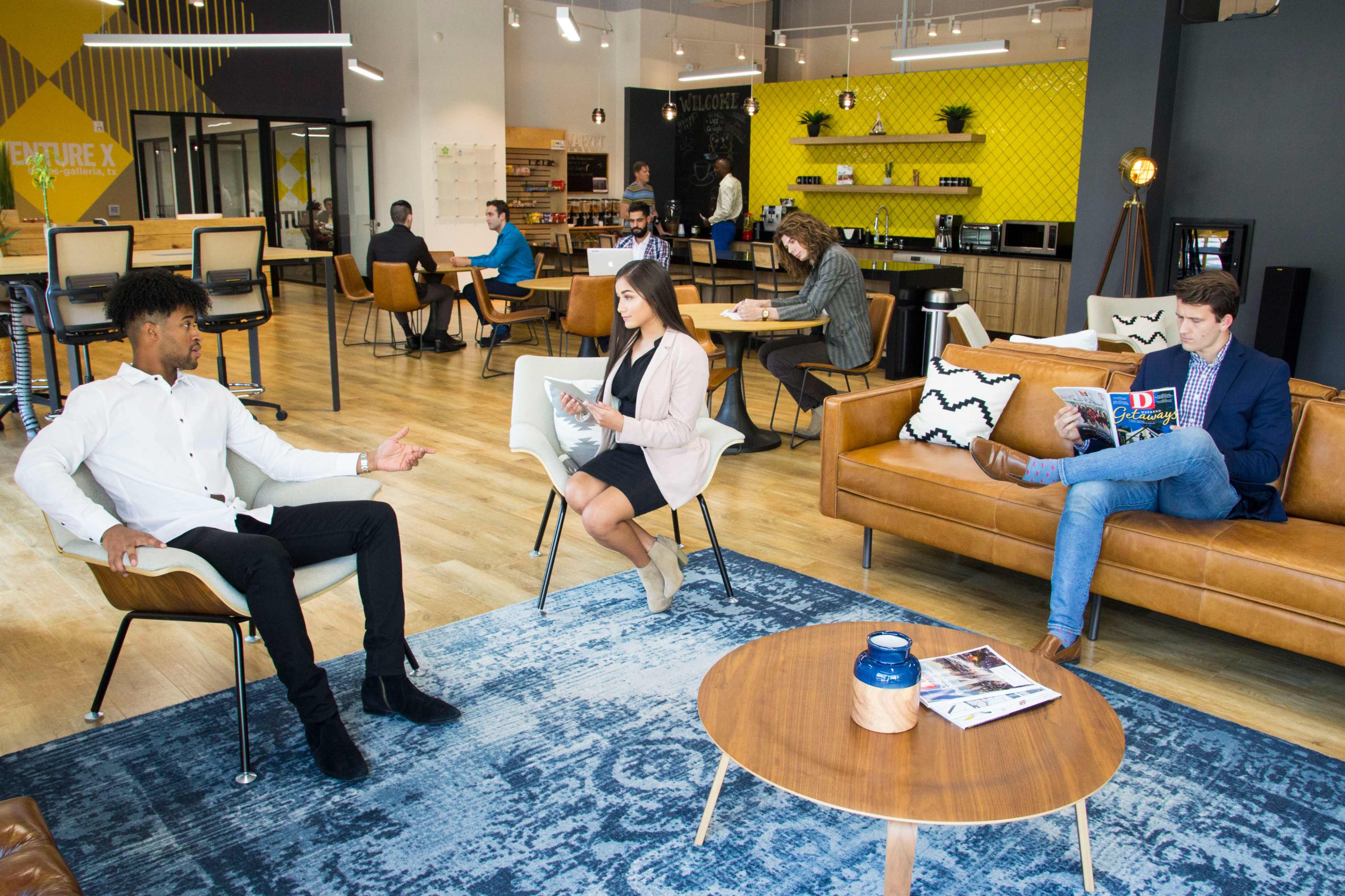 A modern co-working space features several people engaged in various activities, including discussions, reading, and working on laptops, with a relaxed seating arrangement and contemporary decor.