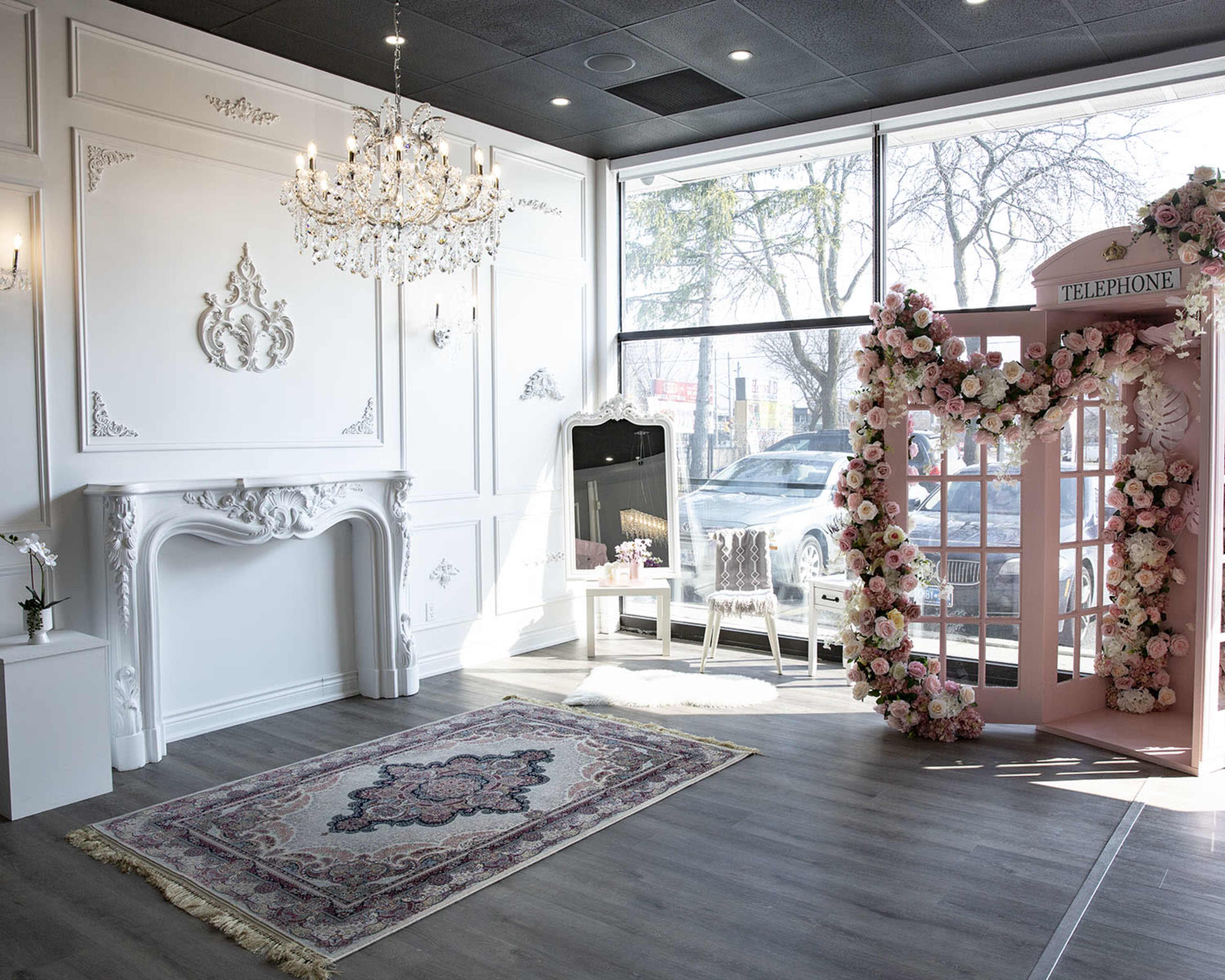 An elegantly designed event space featuring a pink floral archway resembling a vintage telephone booth, with a chandelier and large windows allowing natural light to enter.