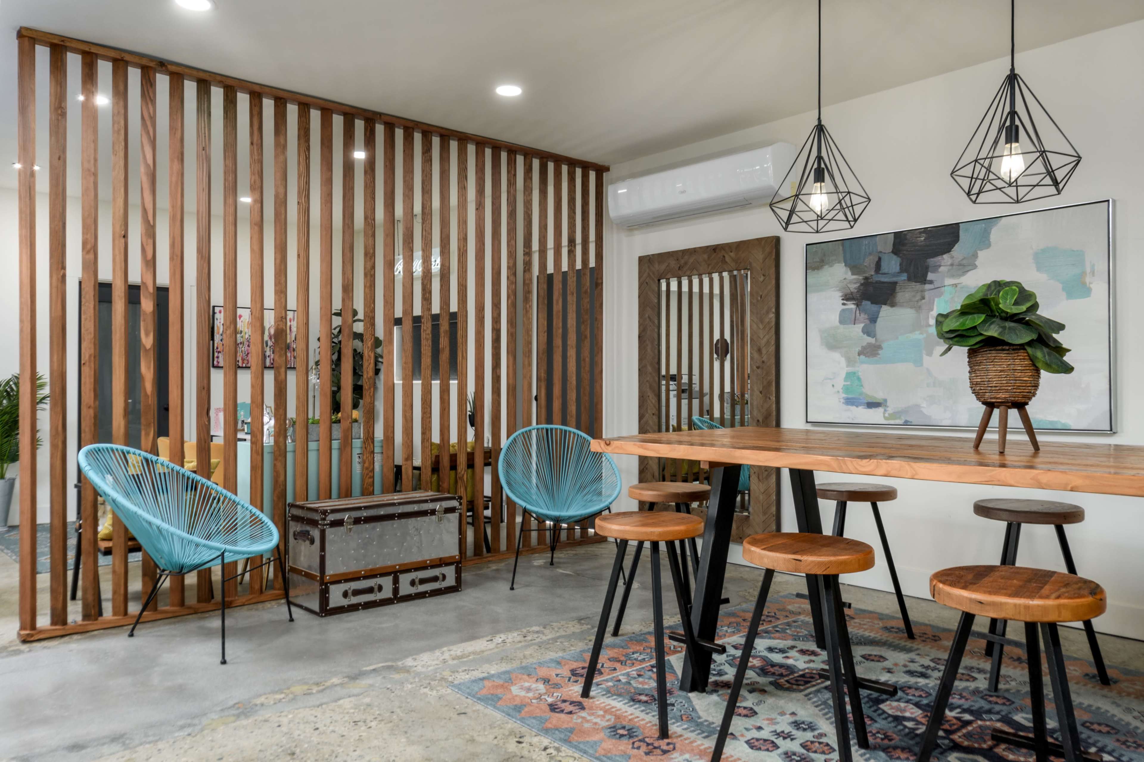 A modern interior features a wooden slatted partition dividing a dining area from a seating space, with vibrant chairs, a wooden table, and hanging pendant lights.